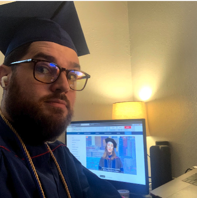 A man in graduation cap and gown sitting in front of a computer monitor with a woman on the screen.