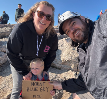 A smiling family of three at Mount Blue Sky, holding a sign with the date 11/20, outdoors with rocks and other people in the background.