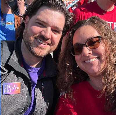 Two smiling people taking a selfie outdoors, with a crowd and bright sunlight in the background.