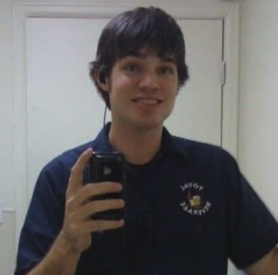 Young man with dark hair taking a selfie in front of a mirror, wearing a blue collared shirt with a logo.