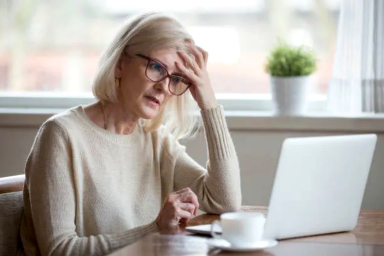 An older woman with glasses sits at a table, looking concerned or stressed while using a laptop.