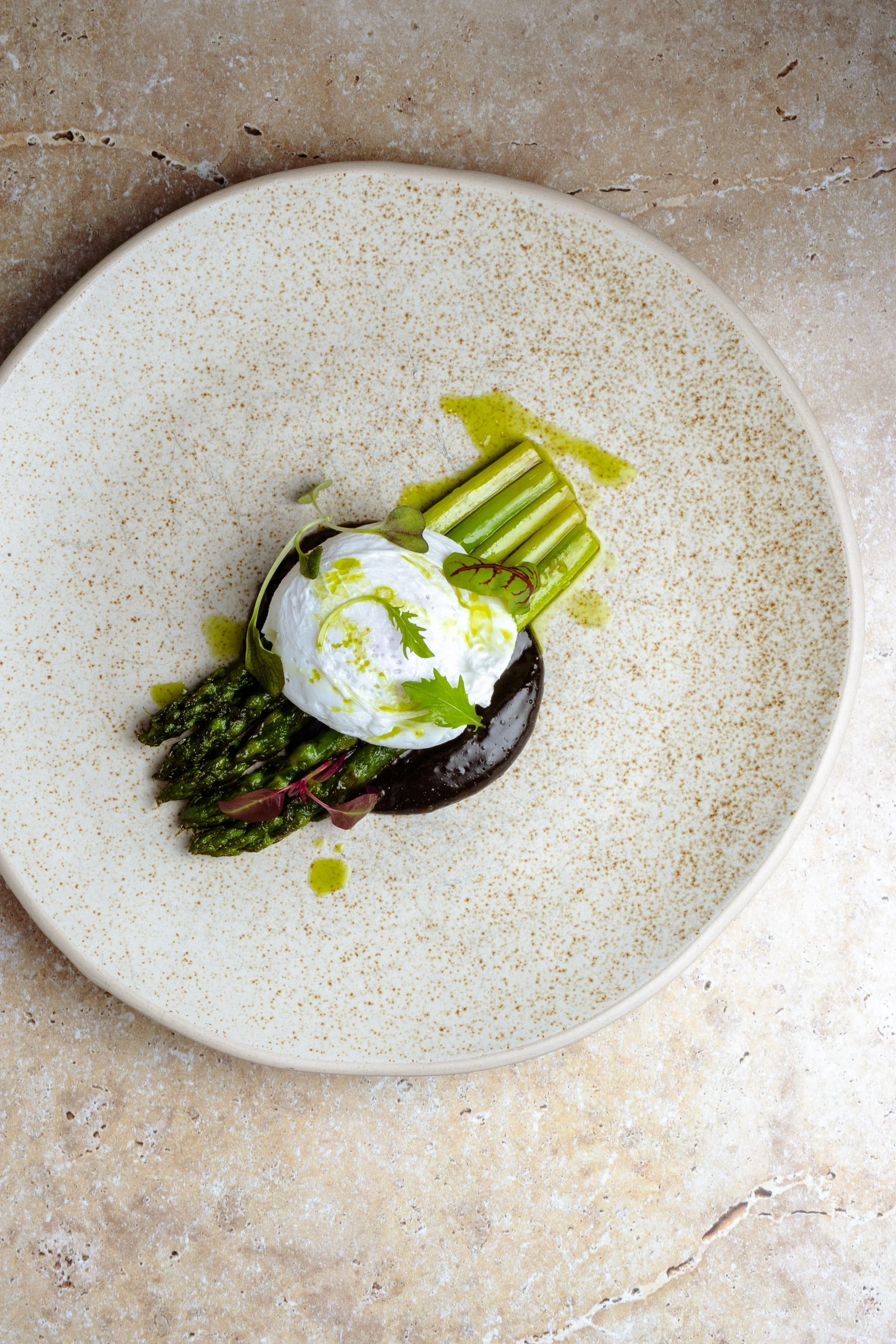 Fresh asparagus spears topped with poached egg, microgreens, and green sauce on a white, speckled plate.