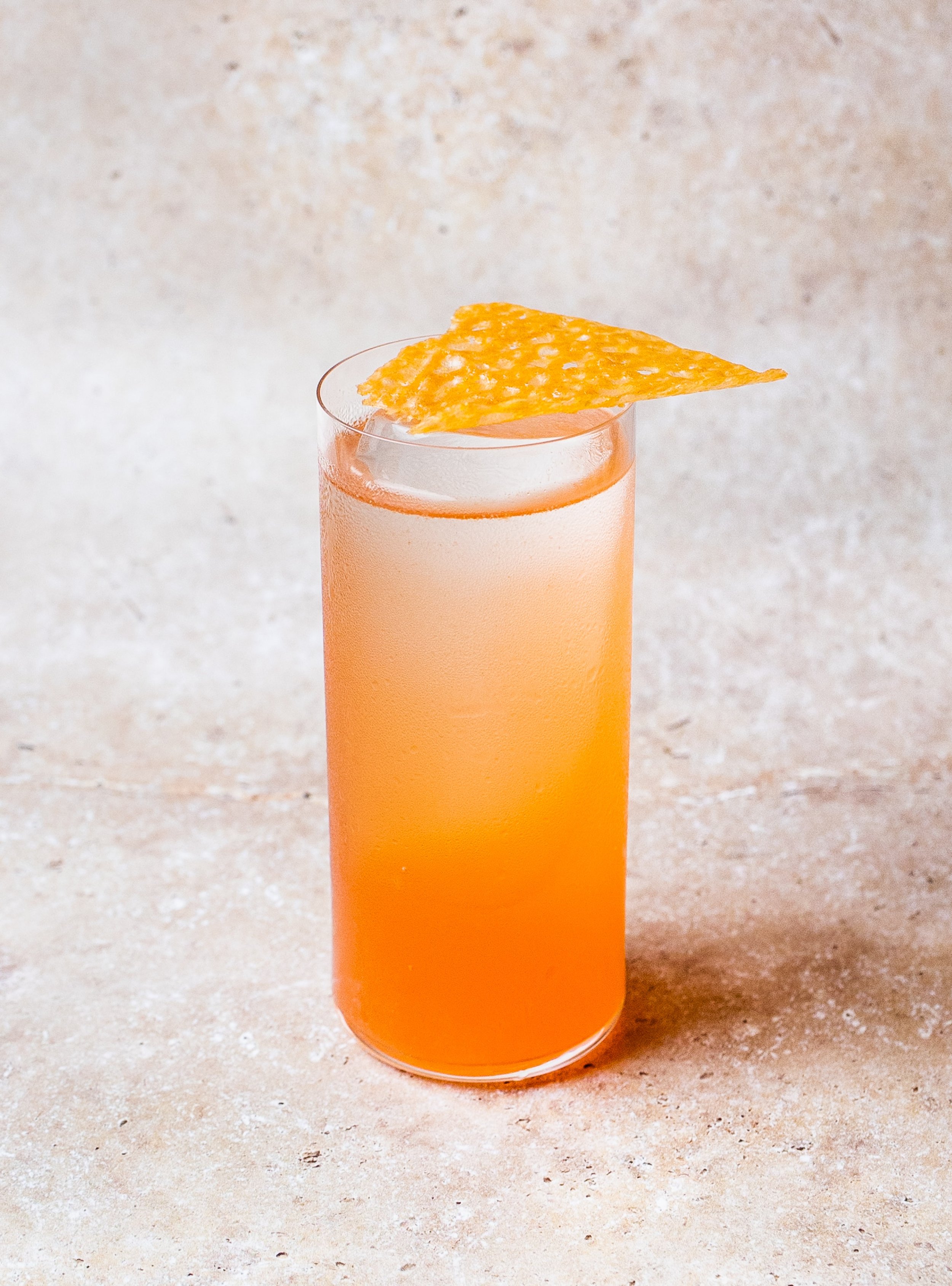 A tall glass of orange-colored cocktail garnished with a crispy, triangular chip on top, placed on a light-colored surface.