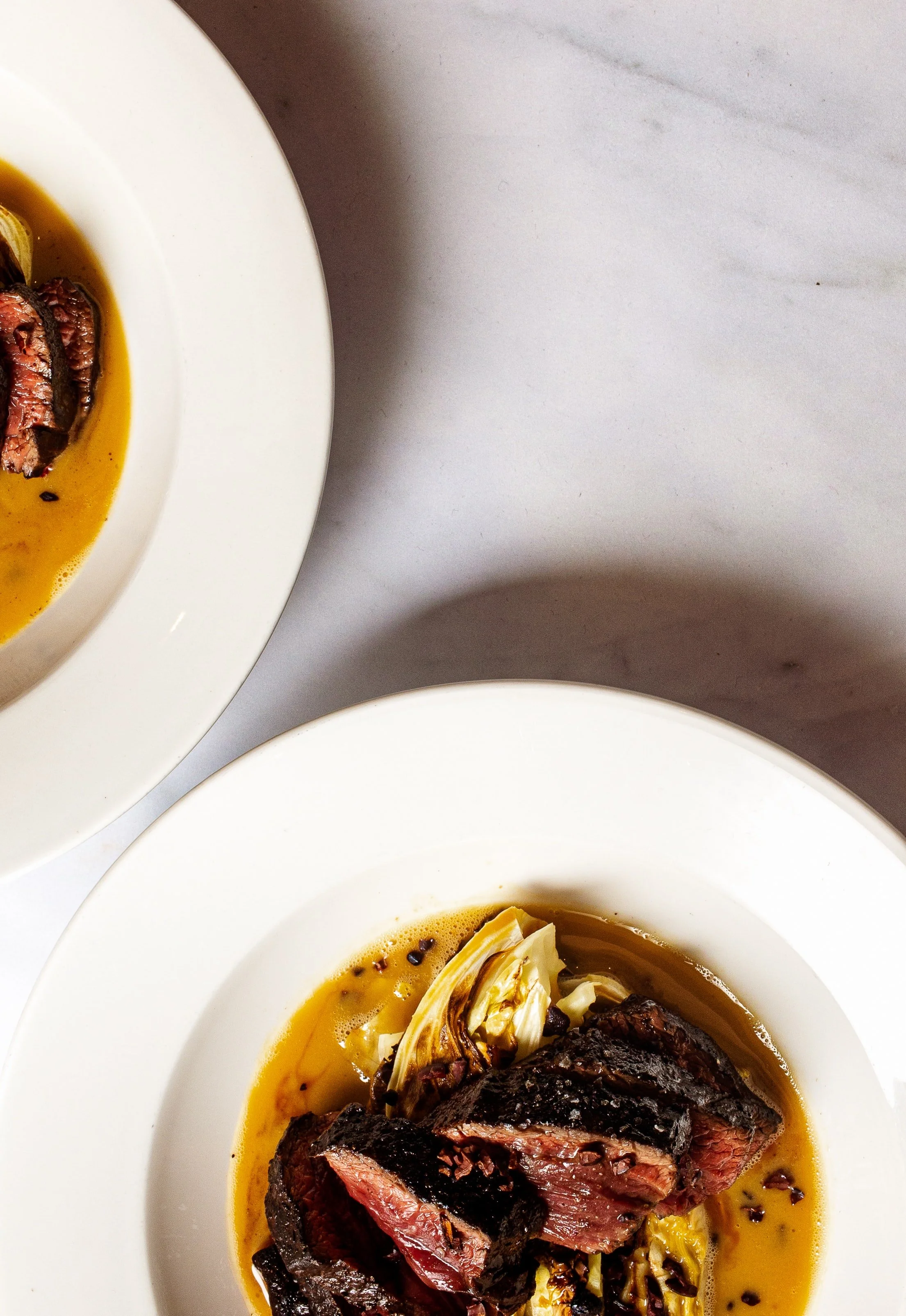 Close-up of two white bowls of cooked meat with sauce, garnished with roasted garlic and black pepper, on a marble surface.