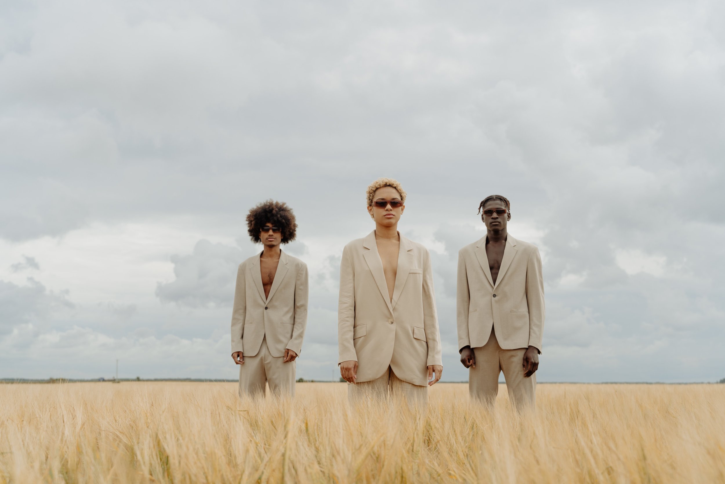 Three people standing in a field of tall golden grass under a cloudy sky, all wearing beige suits and sunglasses.