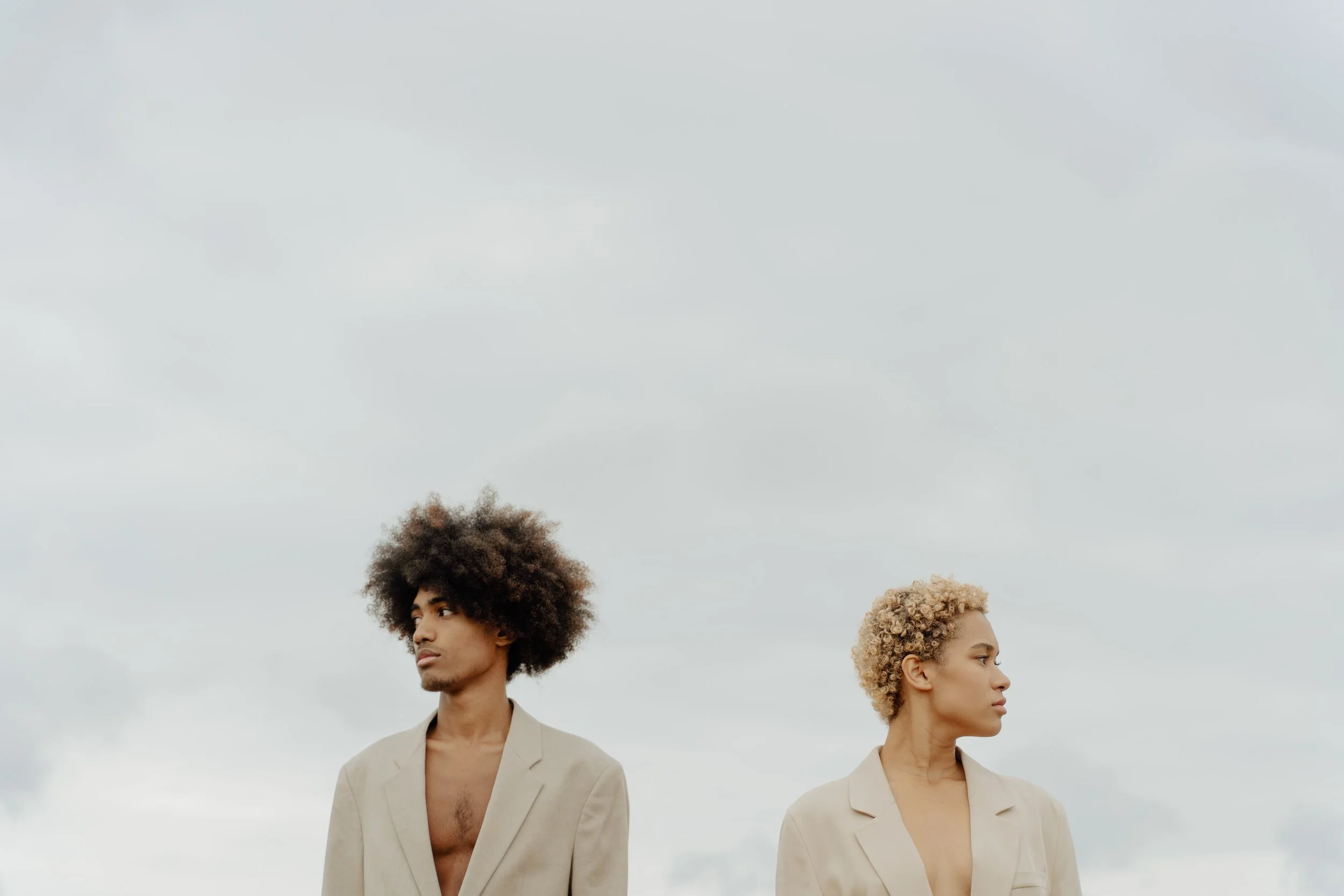 Two individuals standing outdoors on a cloudy day, facing opposite directions. They are dressed in beige blazers, with no shirts underneath visible.