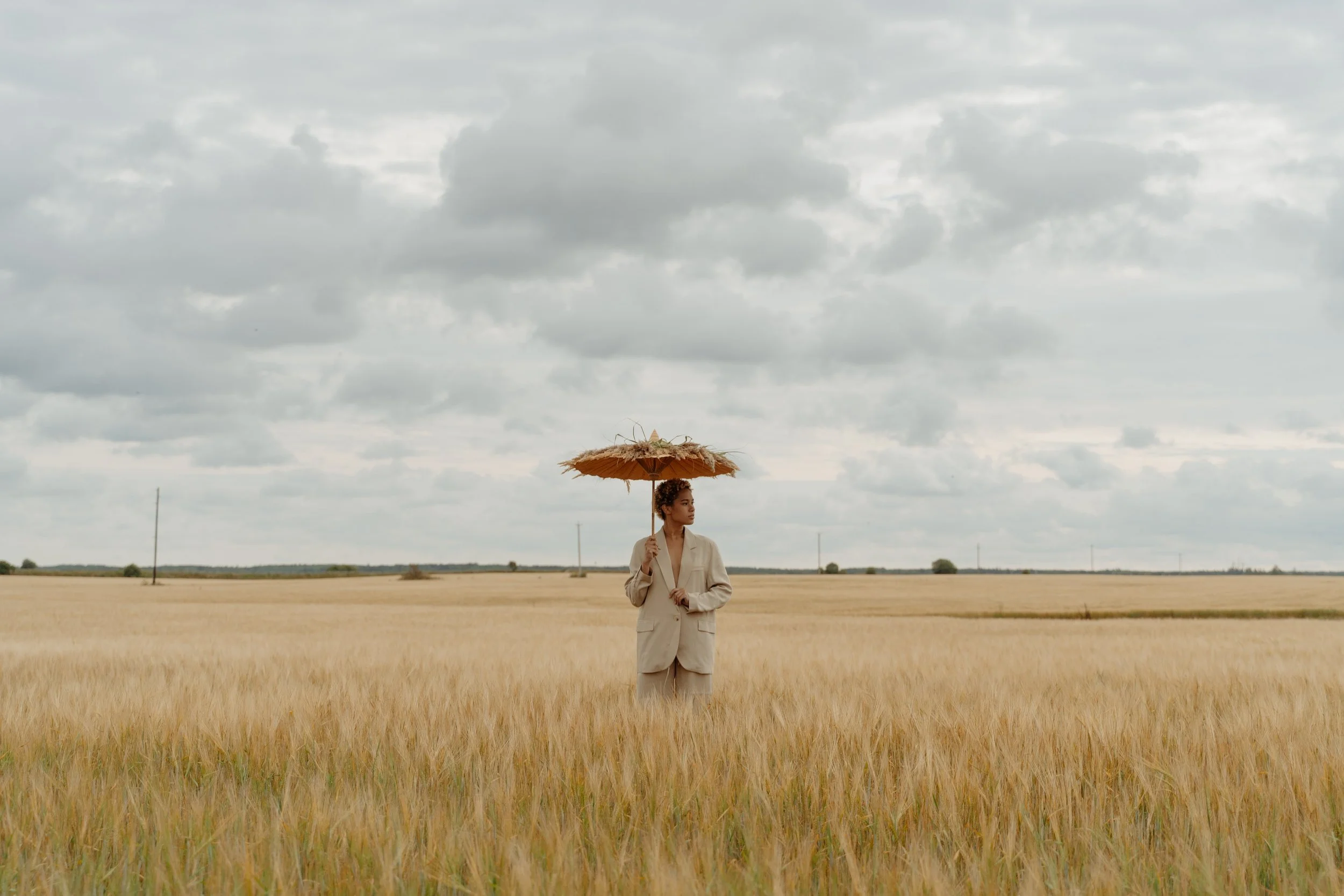 A woman in a beige suit holding a straw parasol stands in a golden wheat field under a cloudy sky.