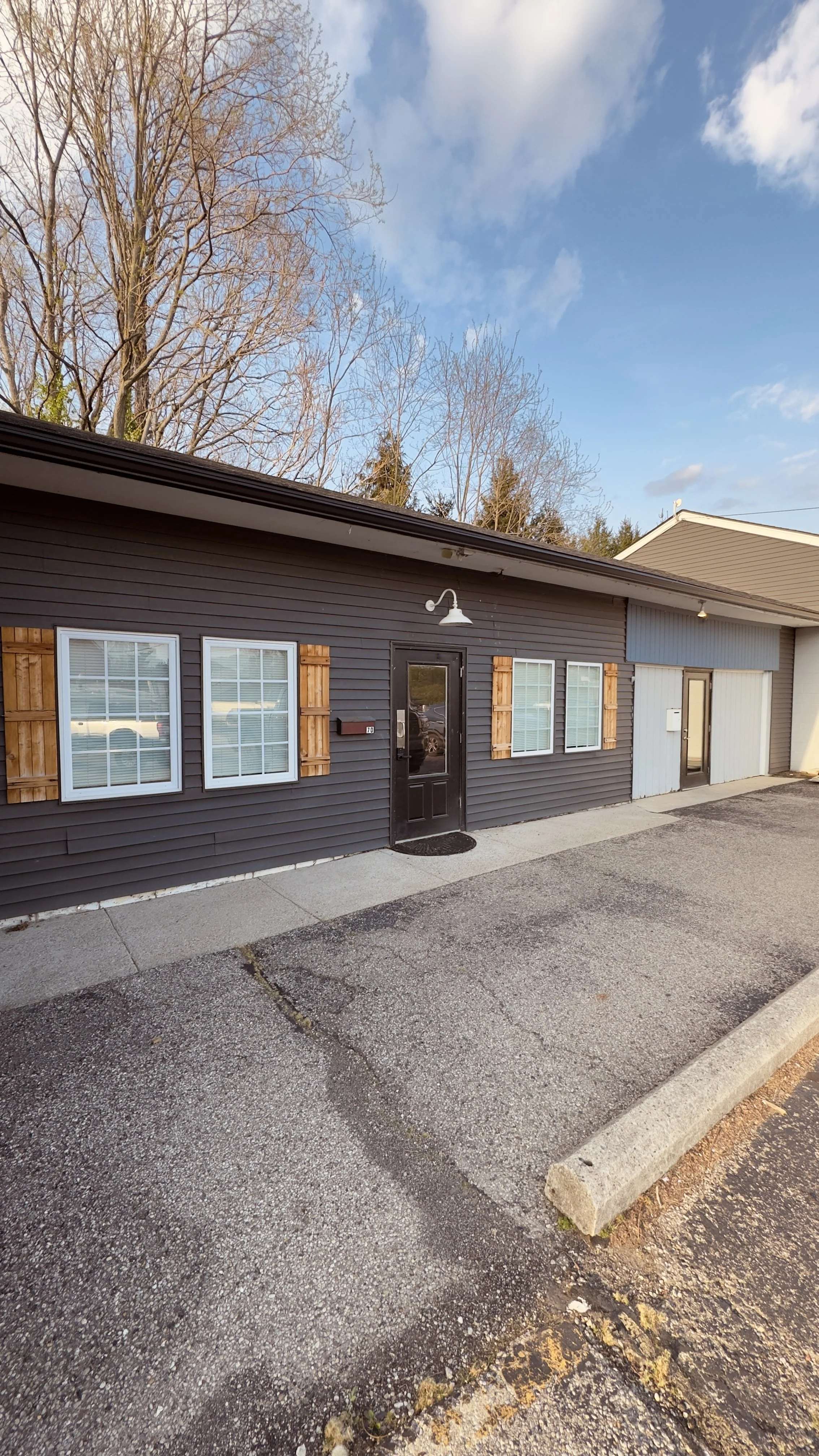 Exterior of a building with dark siding, three windows with wooden shutters, a black door with a white light fixture above, and a parking lot in front under a partly cloudy sky.