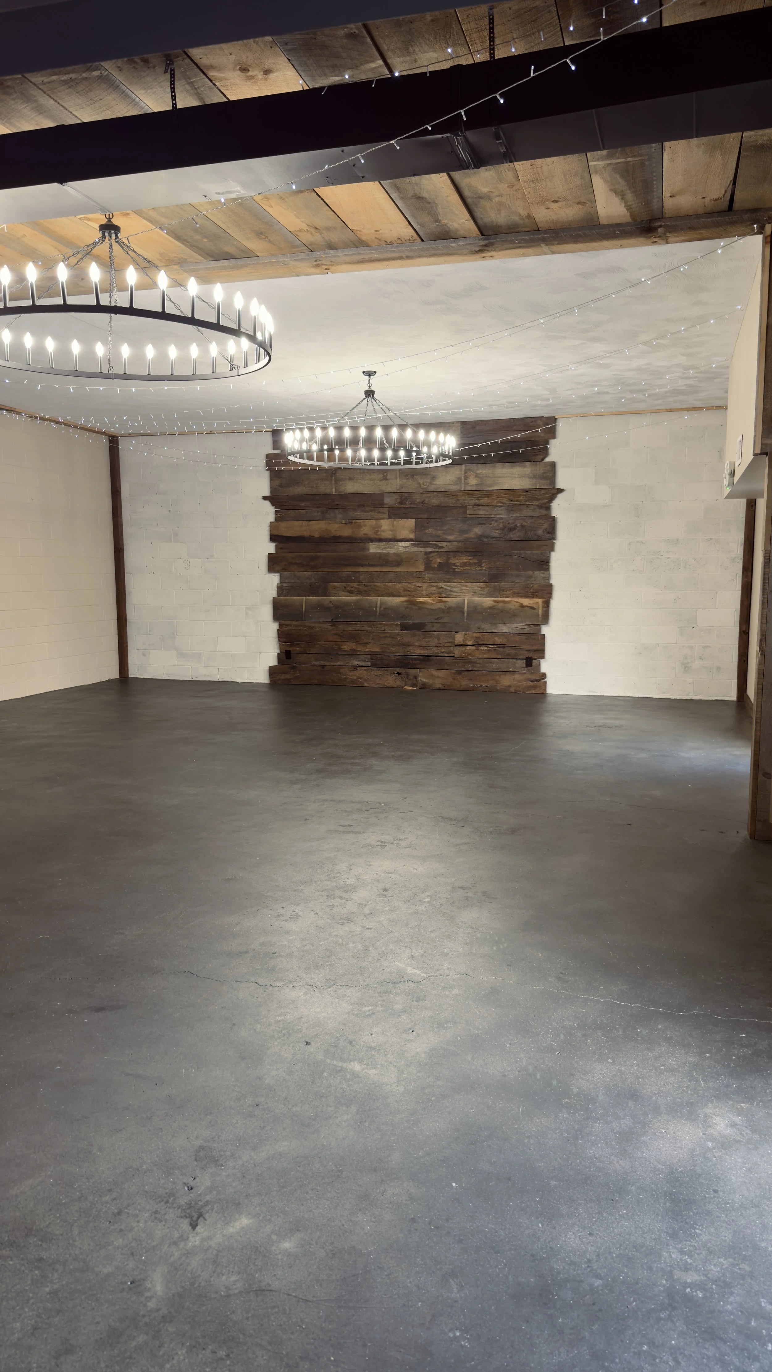 Empty indoor space with stained concrete floor, white brick wall, wooden accent wall, wooden ceiling, two large circular chandeliers, and string lights overhead.