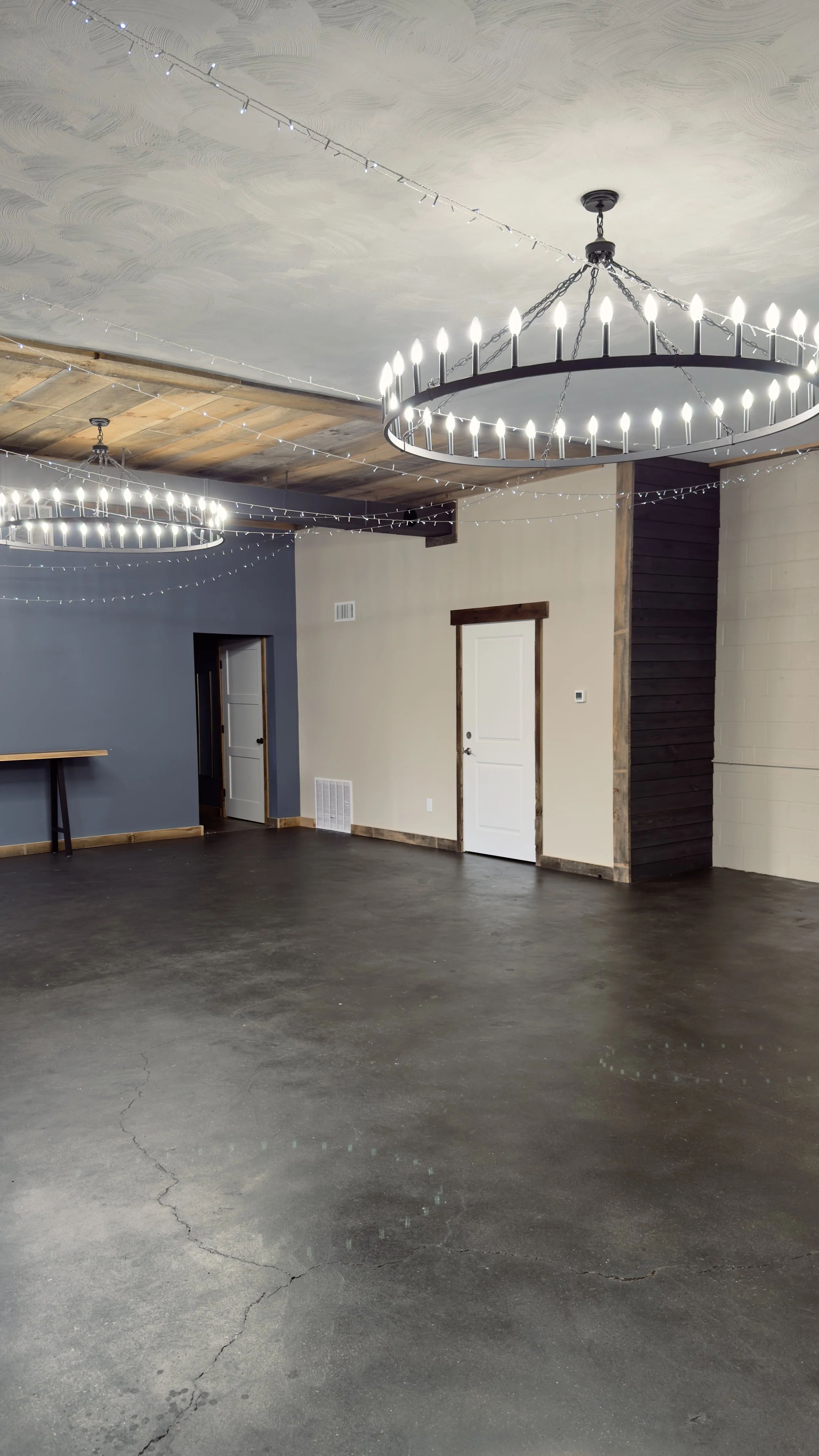 Empty event or party hall with black polished concrete floor, white and blue walls, decorative wooden accents, and two large circular chandeliers with candles, illuminated and hanging from chains. Ceiling decorated with small string lights.