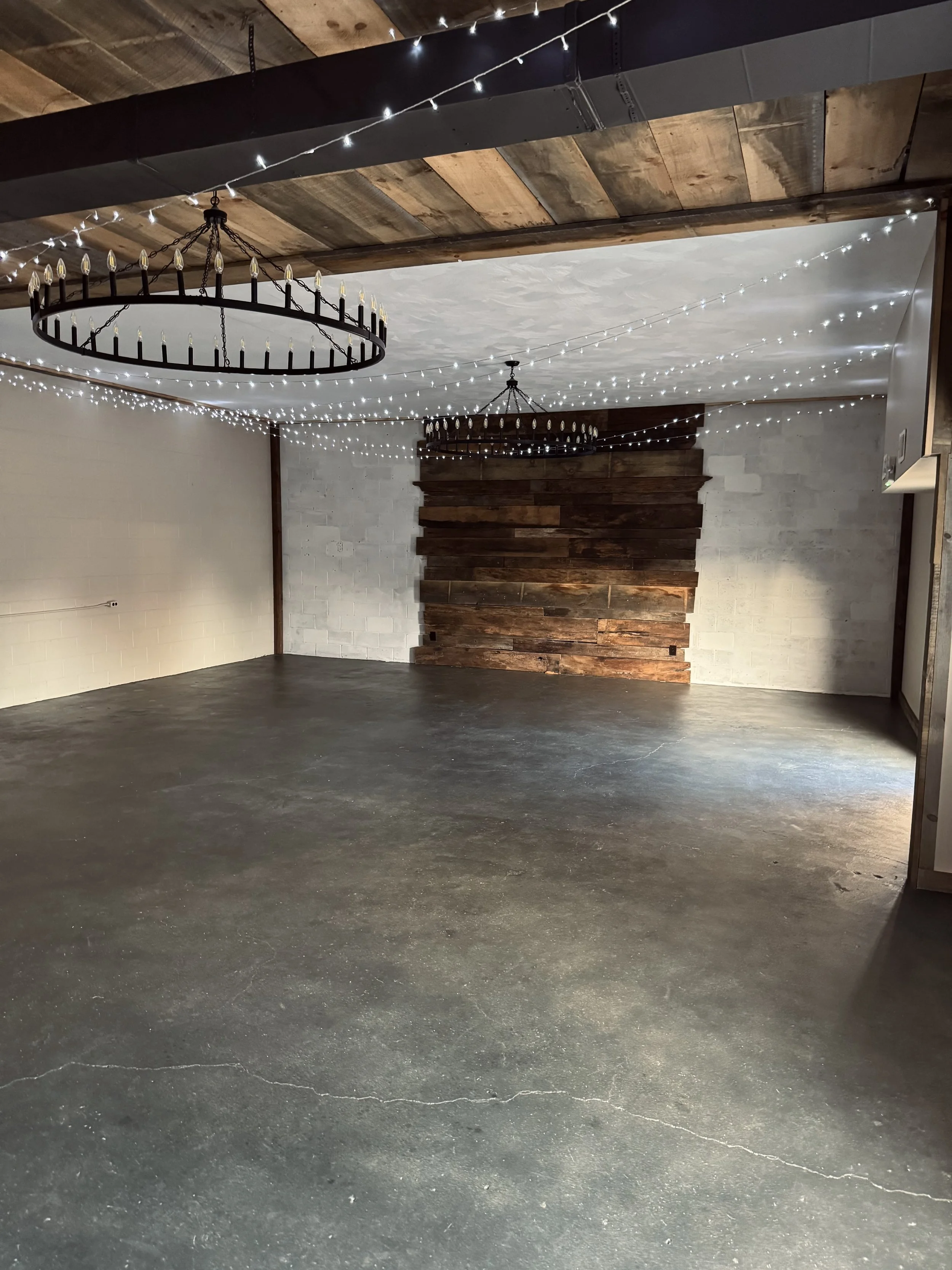 Empty room with string lights hanging from the ceiling, two large circular chandeliers, a wood accent wall, and a concrete floor.