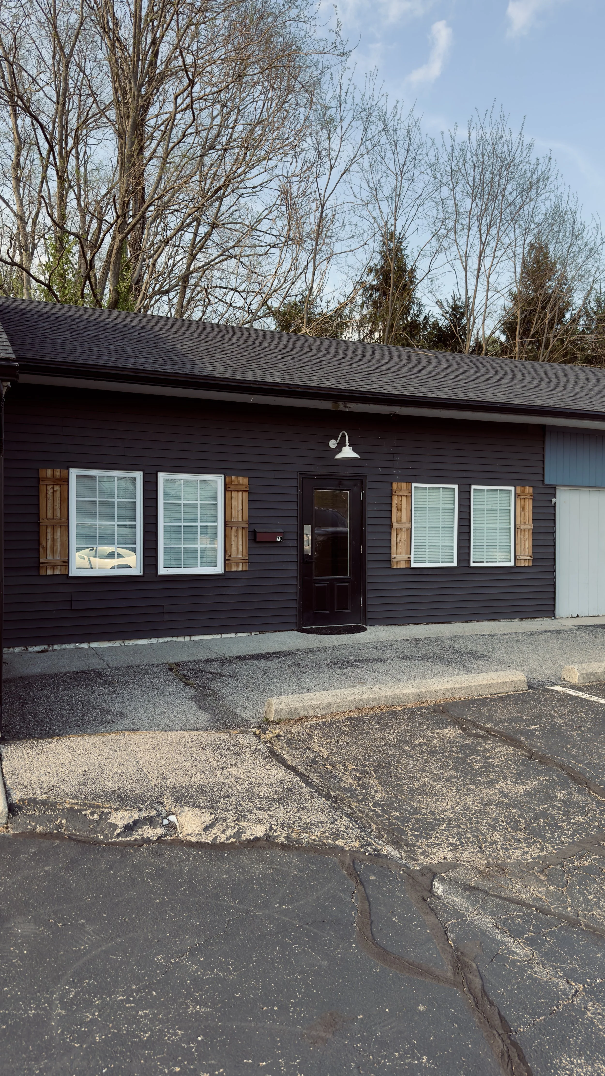 A dark-colored building with three windows and a black door, with a light fixture above the door. The building is set against a background of leafless trees and a blue sky, with a parking lot in front.