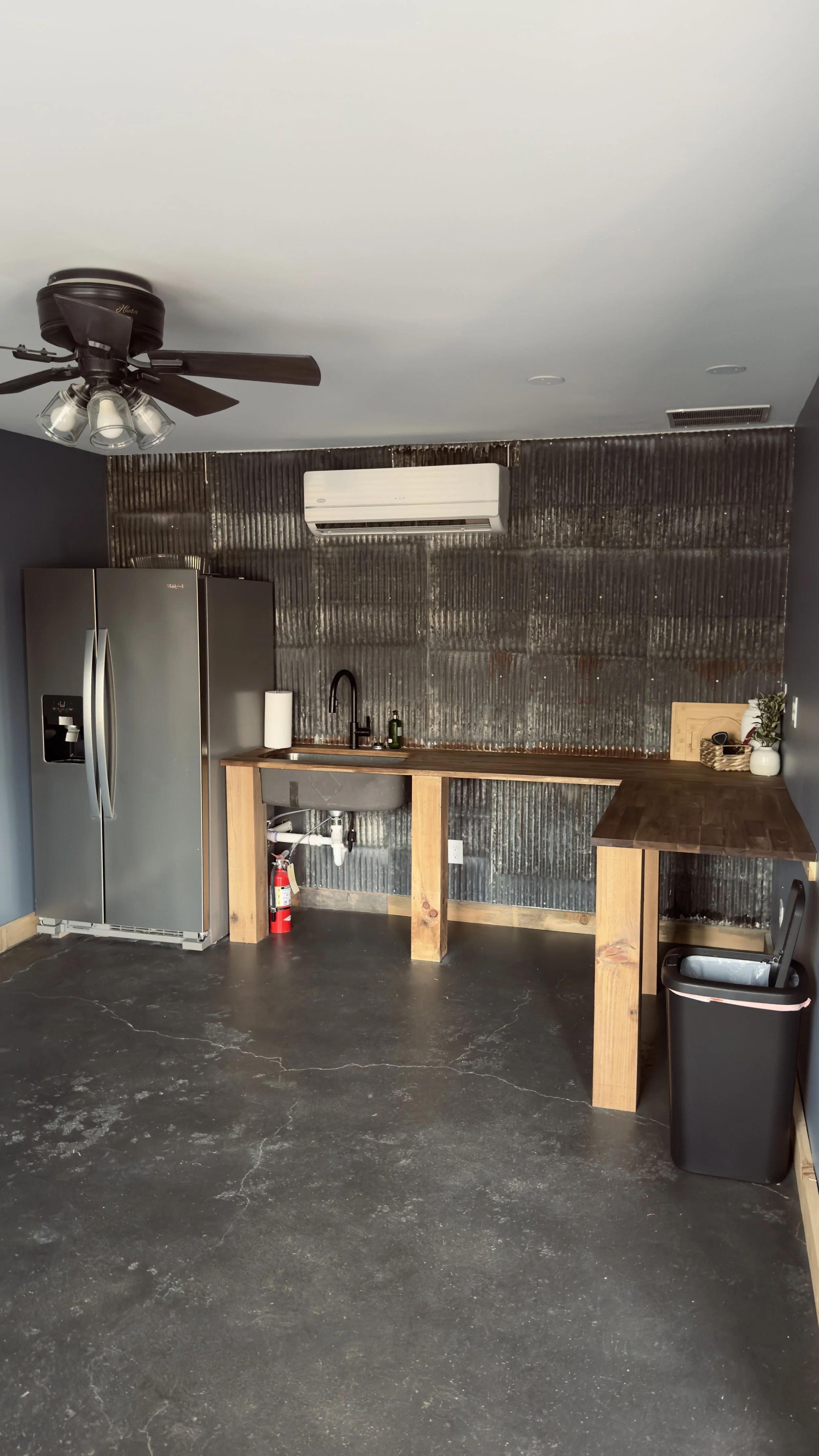 A minimalist kitchen with a stainless steel refrigerator, a black sink with a black faucet, a roll of paper towels, a fire extinguisher, and a wooden countertop against a corrugated metal wall. There is an air conditioning unit on the ceiling and a black trash bin nearby.