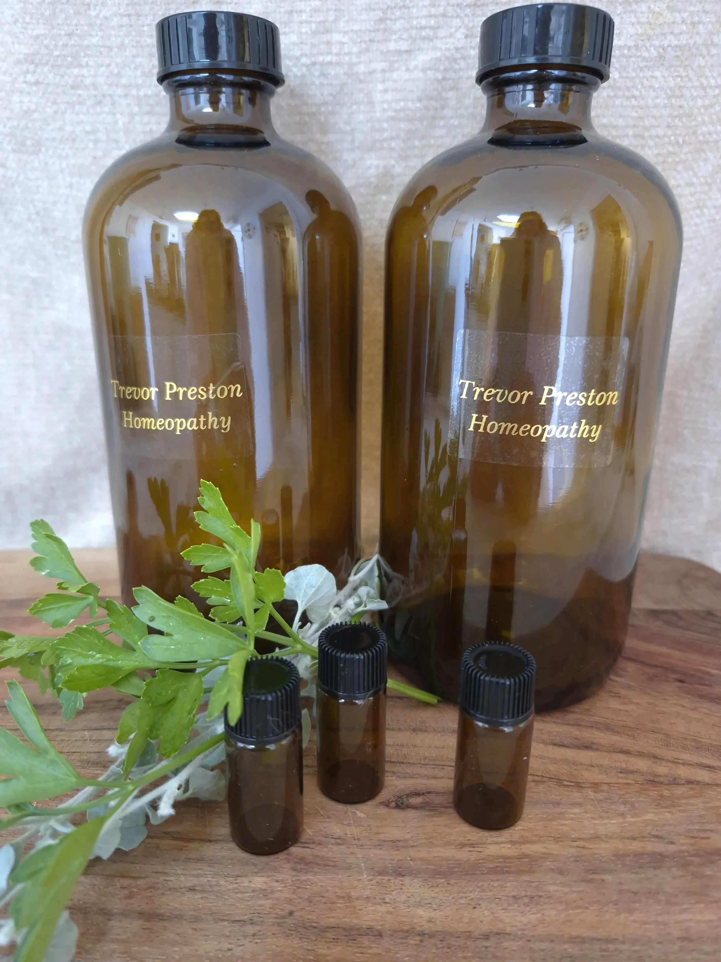 Two amber glass bottles labeled 'Trevor Preston Homeopathy' and three smaller amber glass bottles with black caps, a green leafy plant, and some white plant matter on a wooden surface.