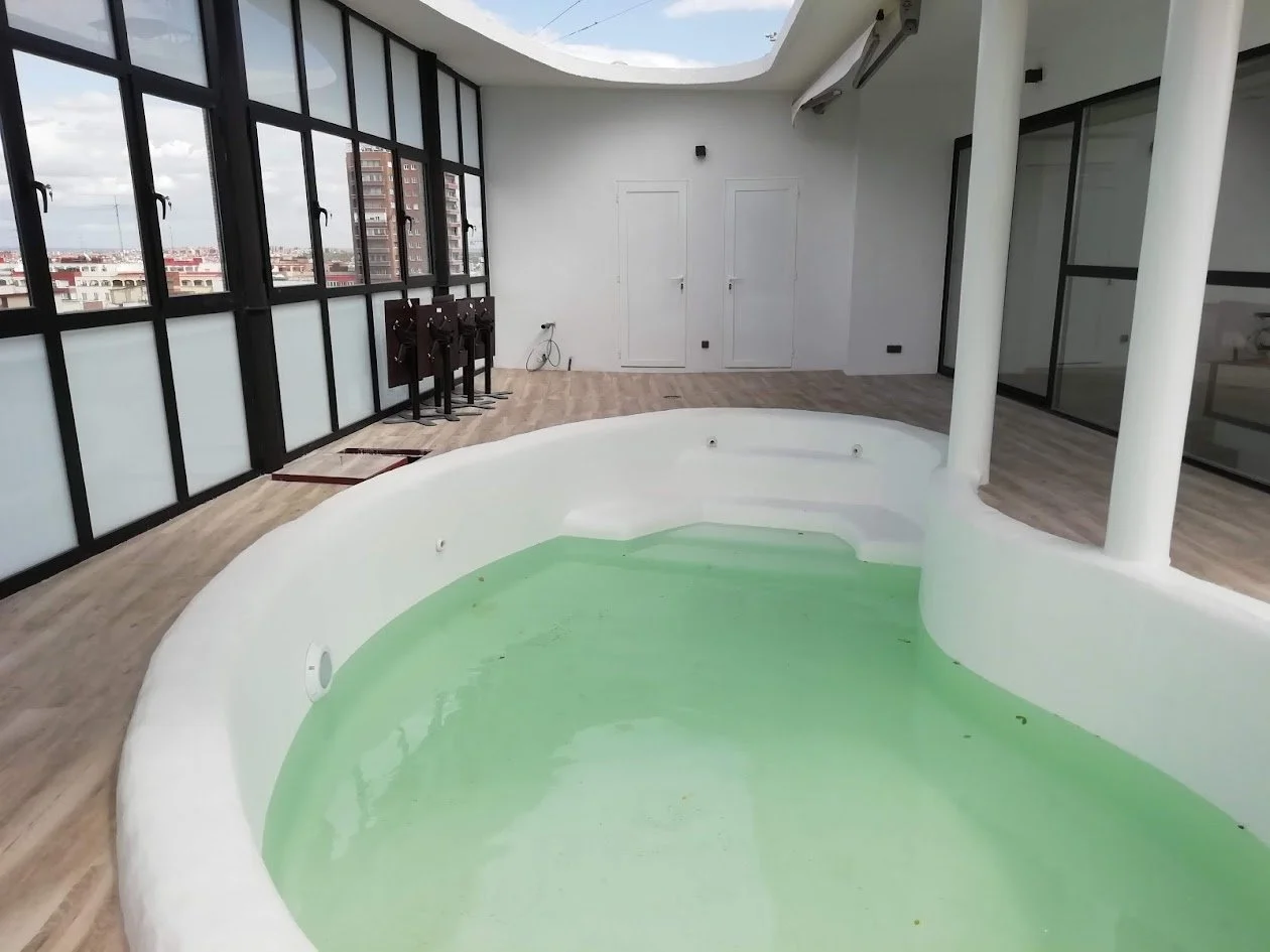 Piscina en un espacio cerrado con ventanas grandes y suelo de madera, con agua de color verde, varias puertas blancas y muebles apilados.