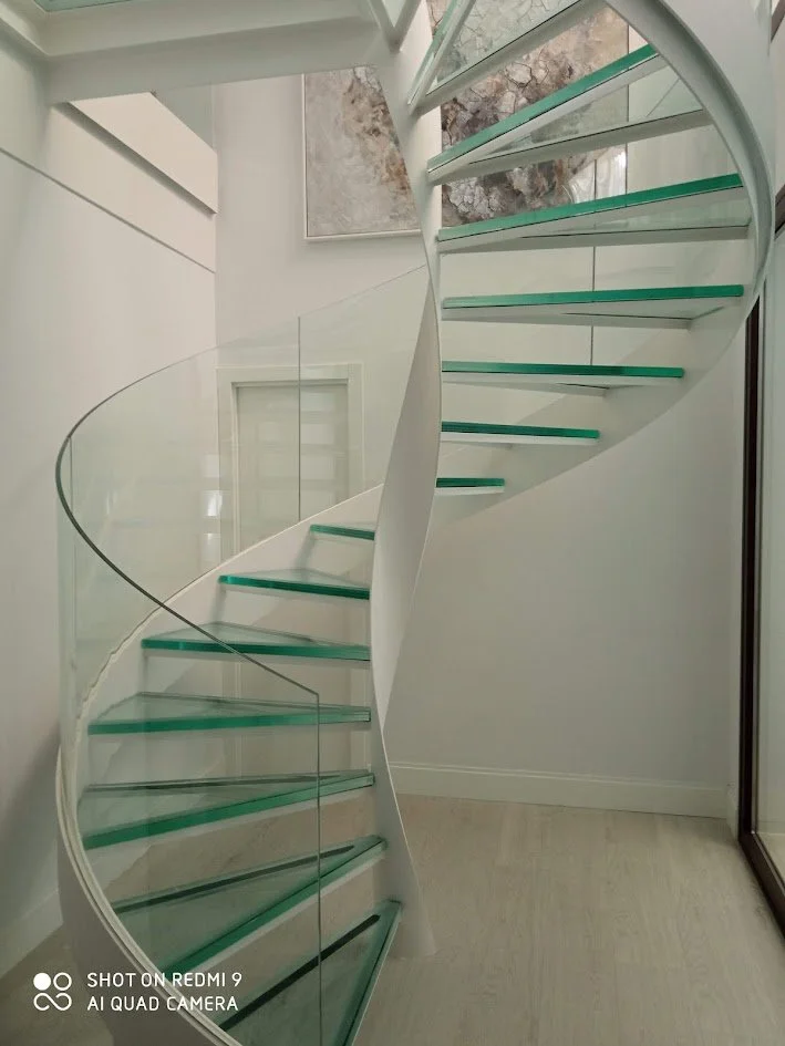 Escalera de espiral moderna con peldaños de vidrio y barandillas de cristal en un espacio interior con paredes blancas y luz natural.