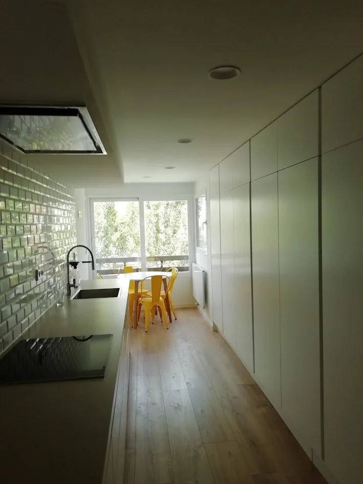 Cocina moderna con encimera, fregadero, estufa eléctrica, armarios blancos, ventanas grandes con vista a árboles, mesa con sillas amarillas, suelo de madera.