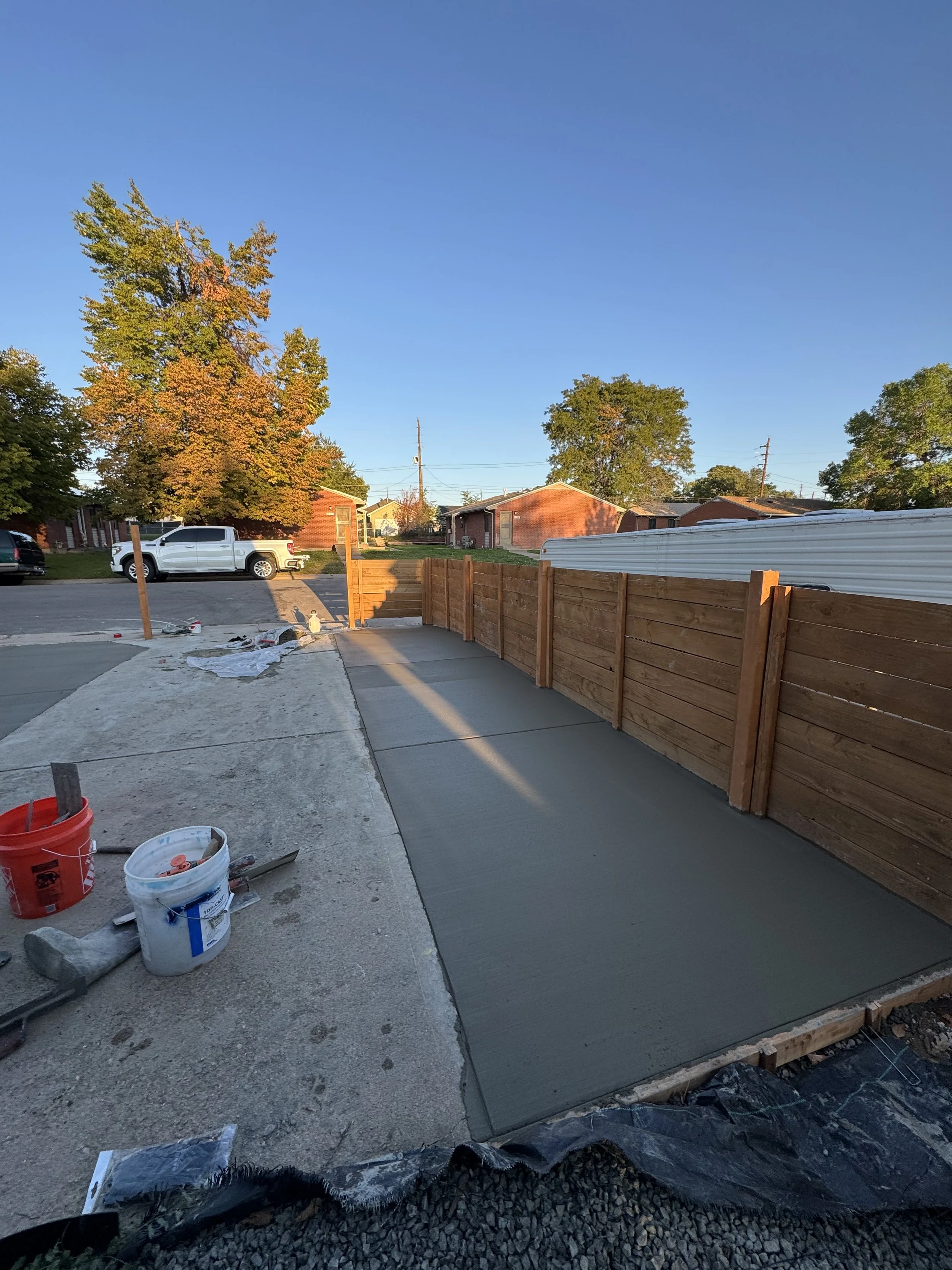 A backyard with a concrete patio, wooden fence, and a freshly poured slab of concrete, with building tools and supplies nearby.