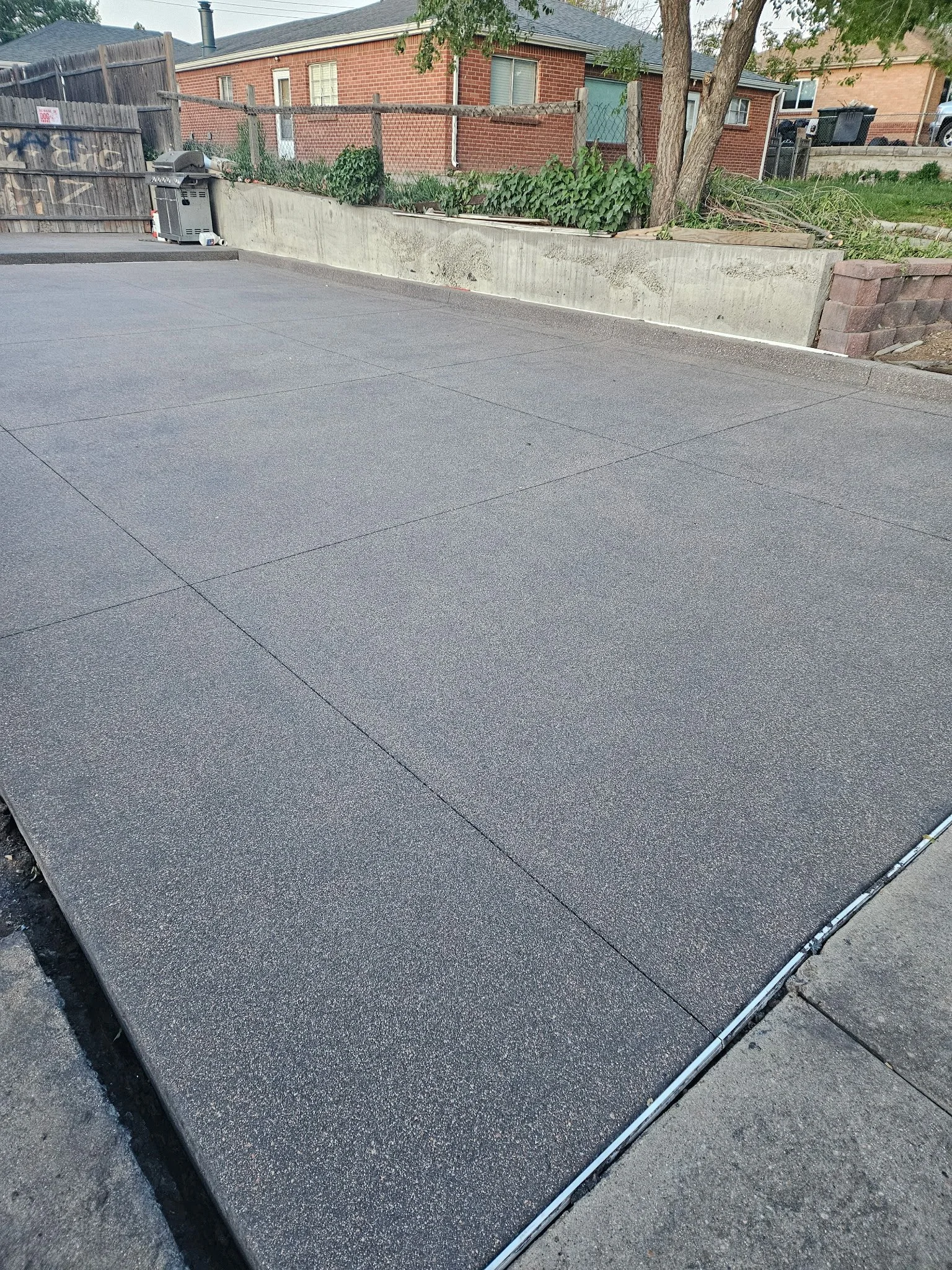Freshly poured asphalt driveway with visible texture and seams, bordered by concrete and brick retaining walls, with residential houses and trees in the background.