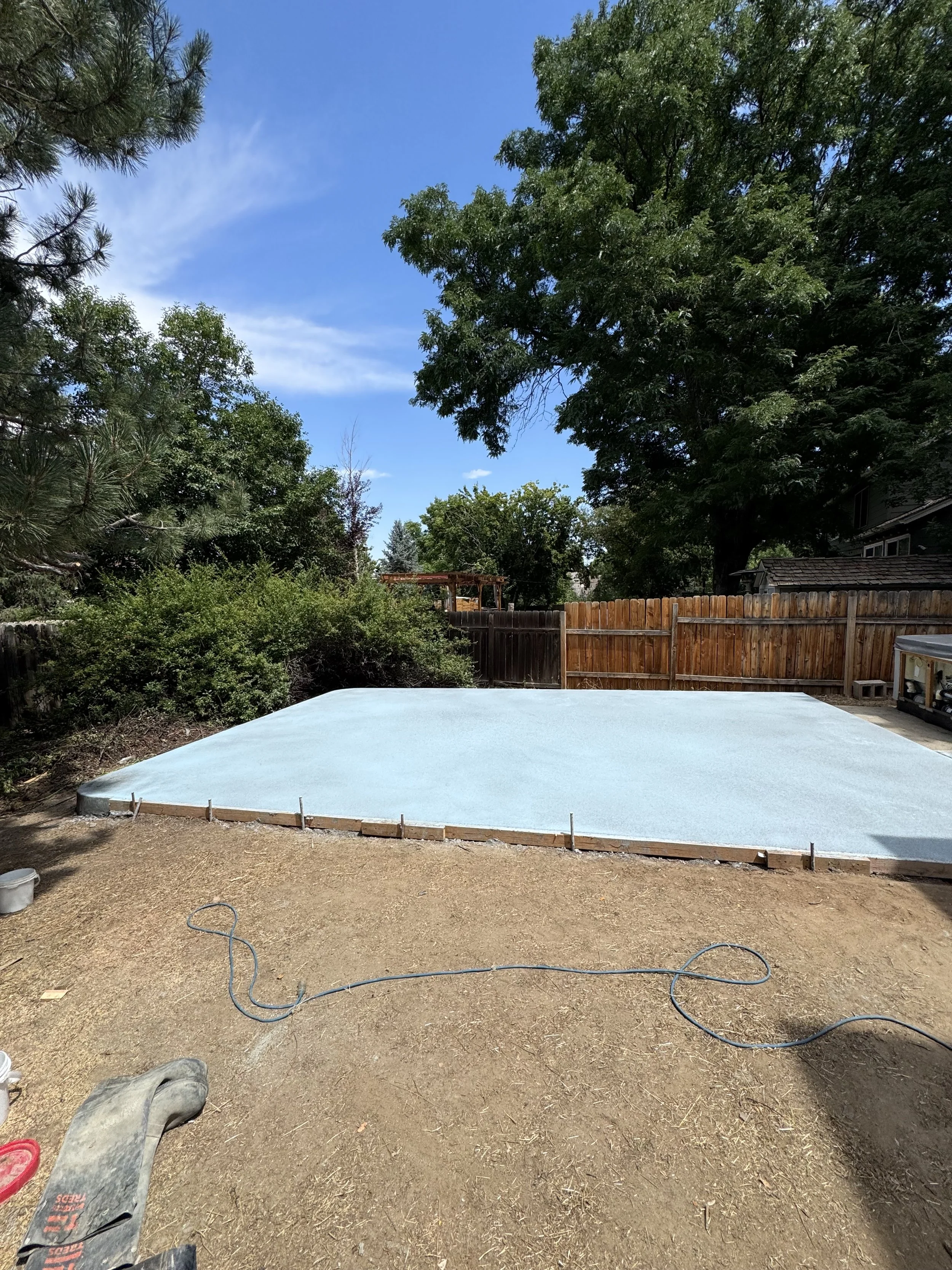 A backyard with a newly poured concrete slab, surrounded by a wooden fence, trees, and bushes under a clear blue sky.