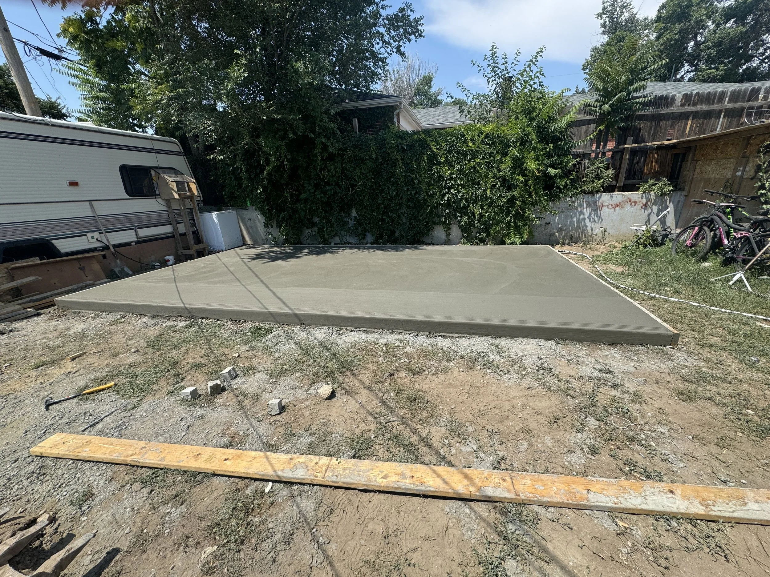 A backyard with an unfinished concrete slab for a patio, construction tools, a trailer, a wooden plank, bikes, trees, and a house in the background.
