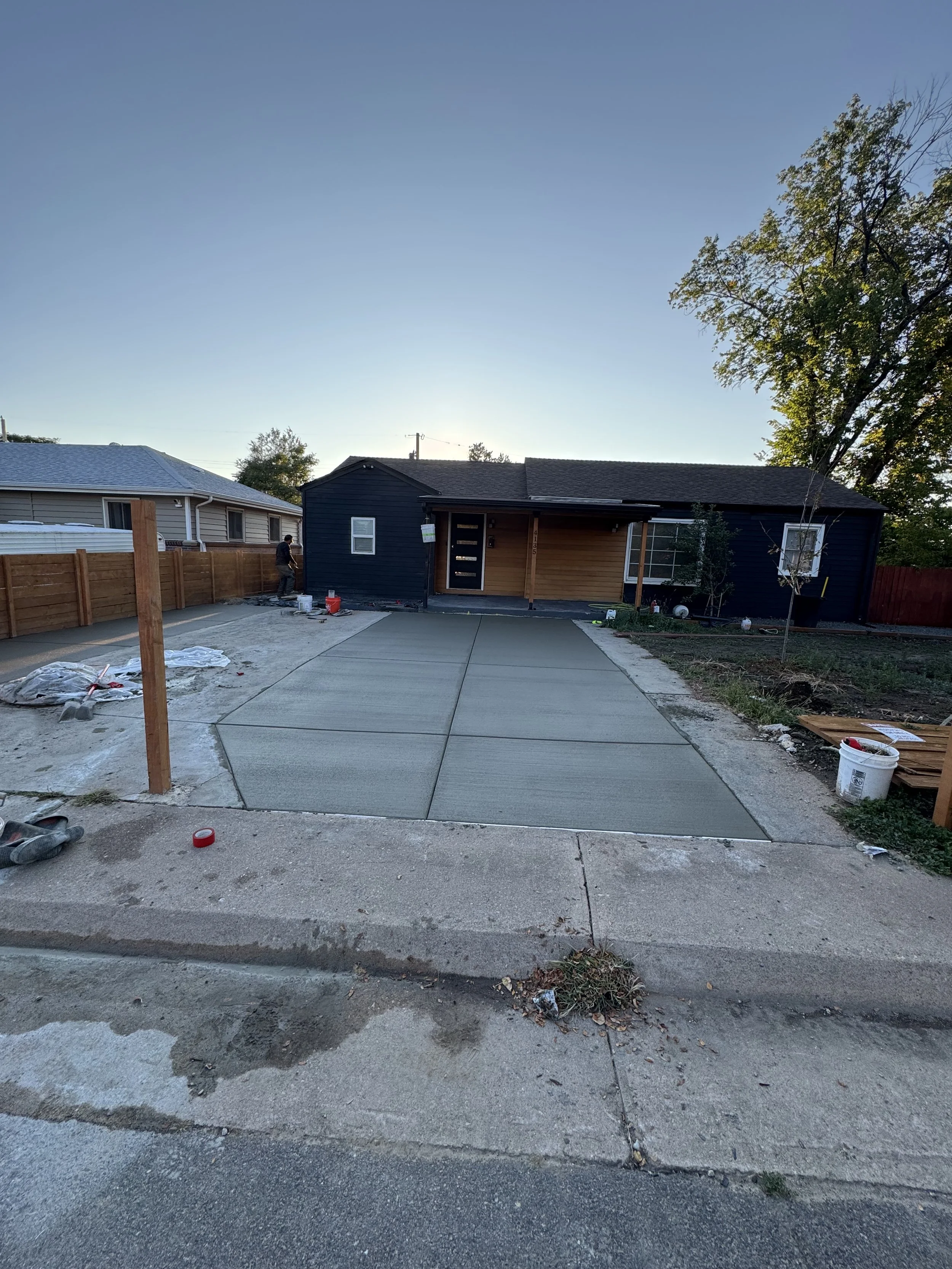 A house under construction with a new concrete driveway, a person working near the house, and construction materials and tools on the ground.
