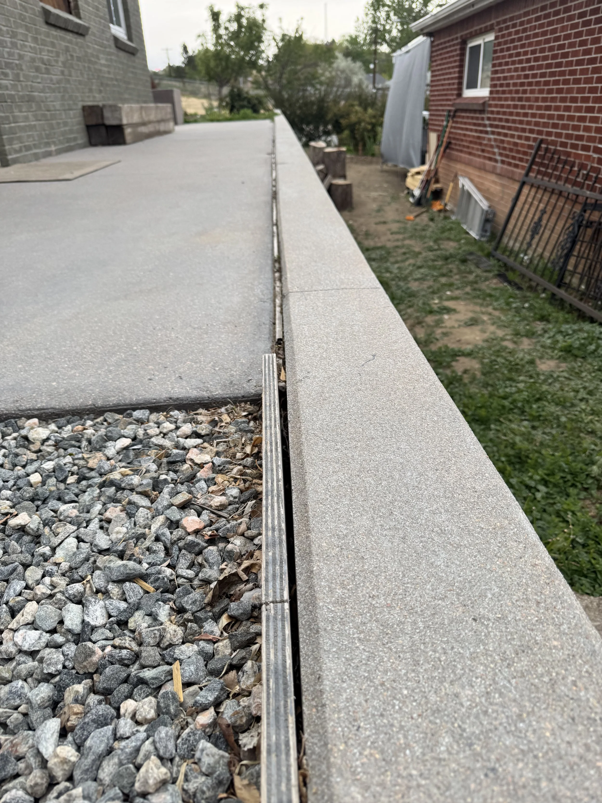 Close-up of a concrete porch with a metal linear drain, with a small section of gravel and rocks underneath and part of a house with brick walls visible on the sides.