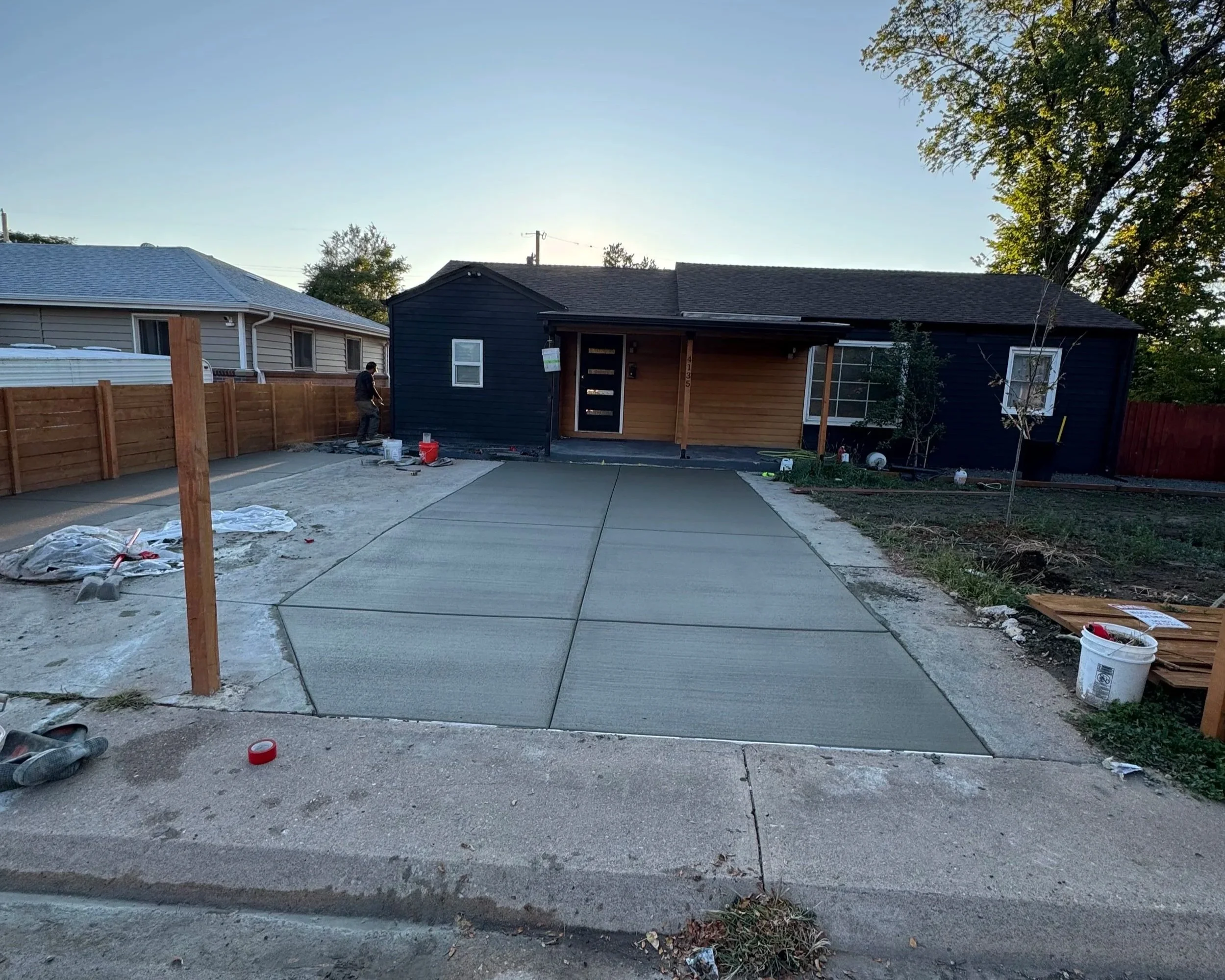 Residential backyard with driveway, wood fence, and two houses under renovation, one blue and one beige, with construction tools and materials scattered around.