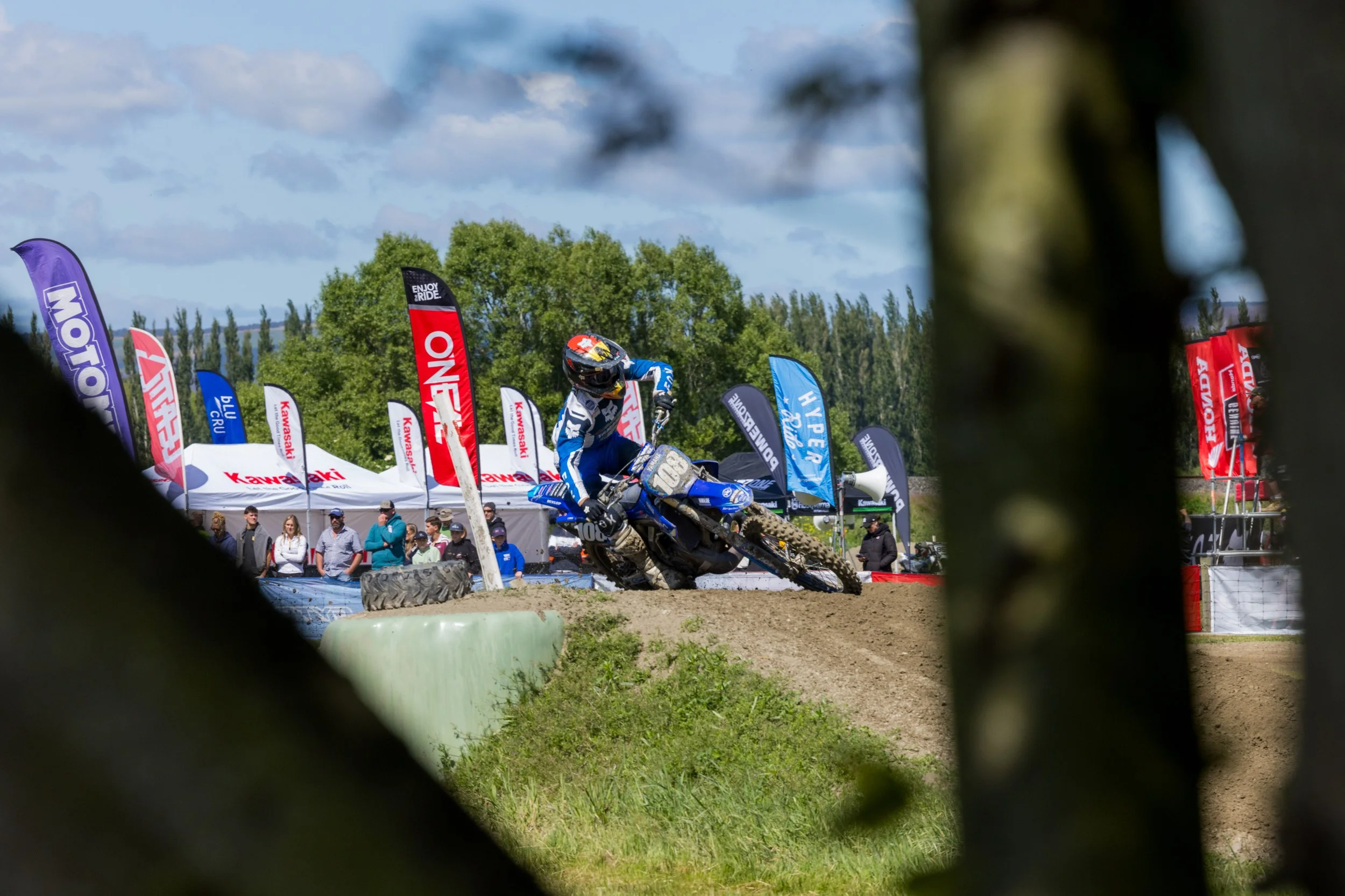 A motocross rider on a blue dirt bike, wearing a helmet and gear, racing on a dirt track near colorful flags and spectators, with trees and a cloudy sky in the background.