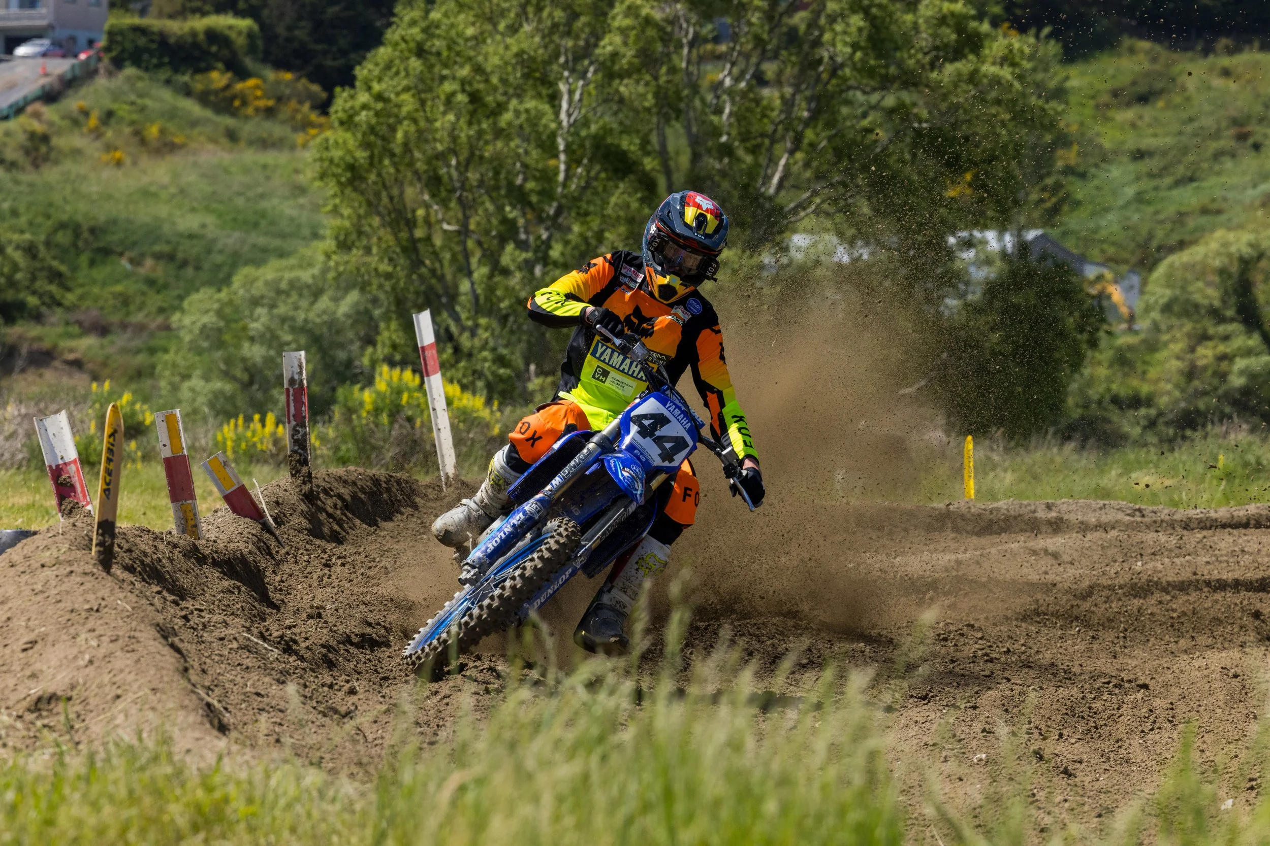 A motocross rider in colorful gear and helmet riding a dirt bike through a dusty track lined with safety poles, with trees and a hillside in the background.