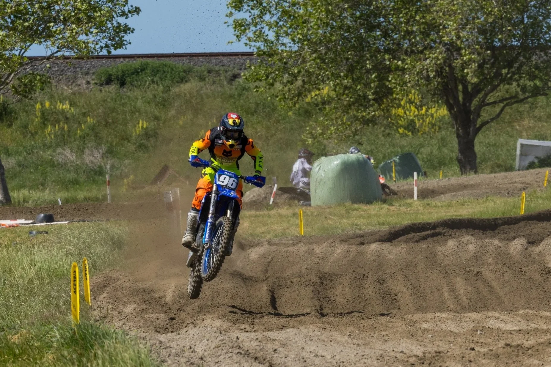 A motocross rider wearing an orange and black suit, helmet, and goggles riding a blue dirt bike with the number 96 on a dirt track, kicking up dust as he speeds around the curve, with green trees and a blue sky in the background.