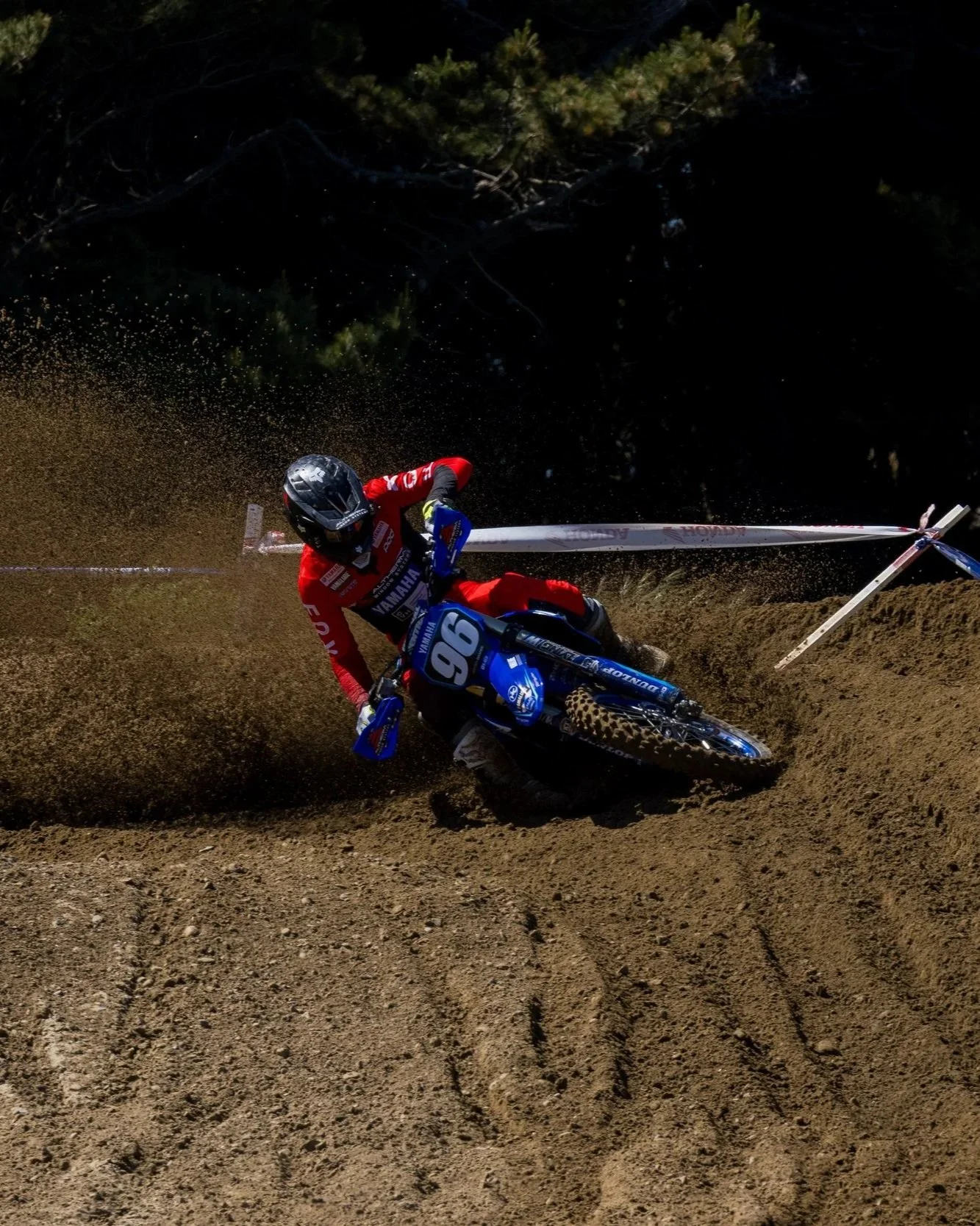 A motocross rider wearing a red suit and black helmet navigating a dirt track, kicking up dirt behind the bike.