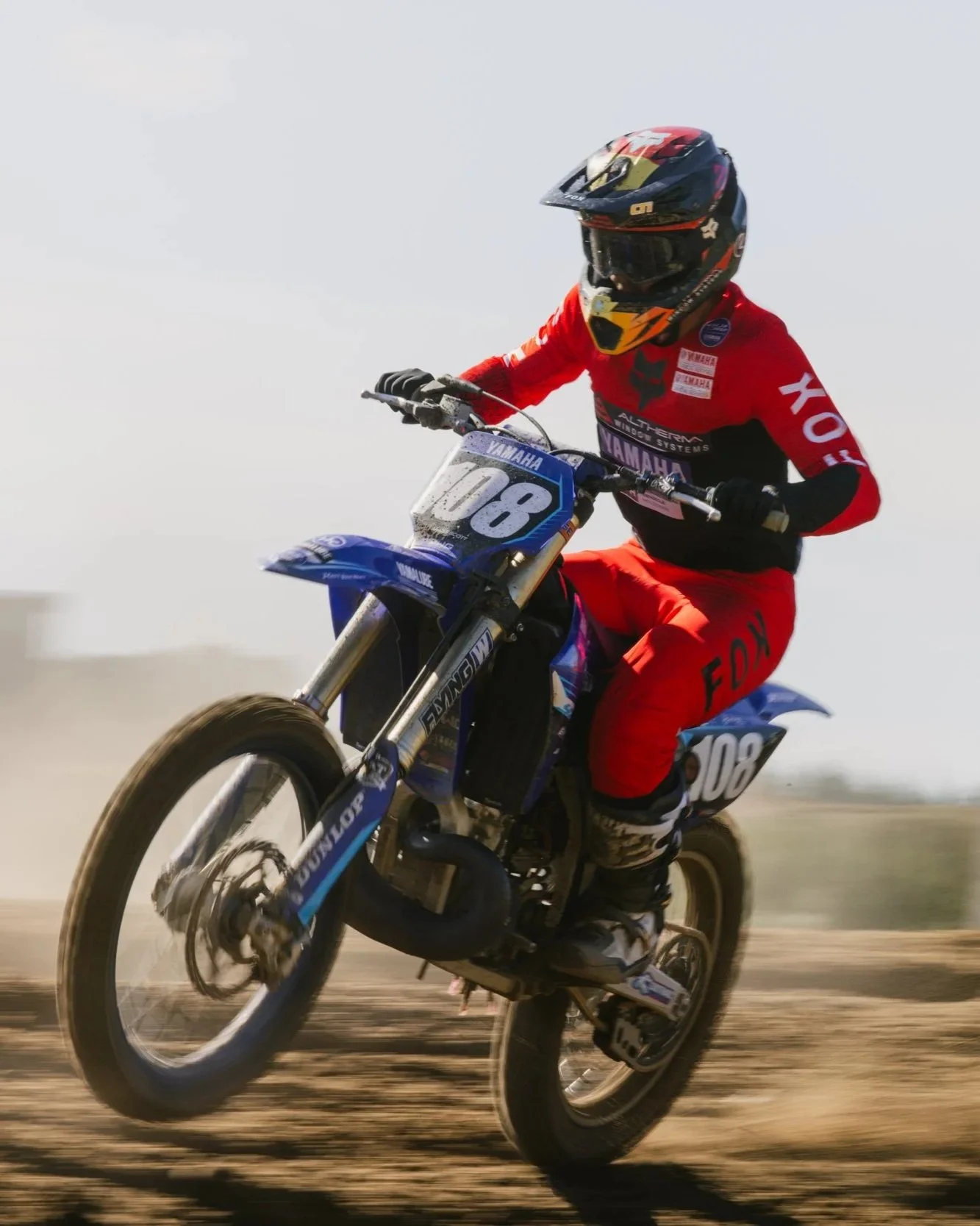 A motocross rider in a red and black suit and helmet racing a blue Yamaha dirt bike on a dirt track.