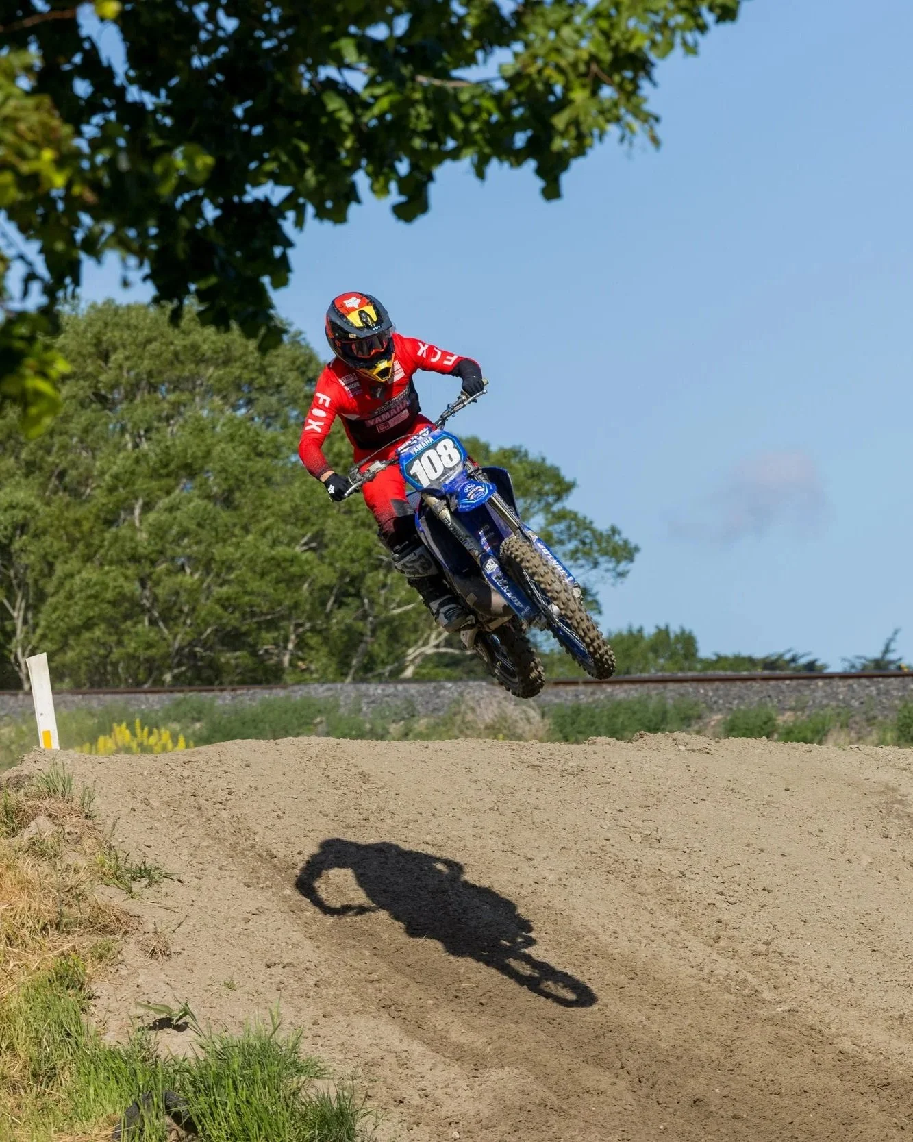 A motocross rider wearing a red and black outfit, helmet, and goggles jumping a dirt mound on a blue motorcycle with the number 108, during daytime with green trees and blue sky in the background.