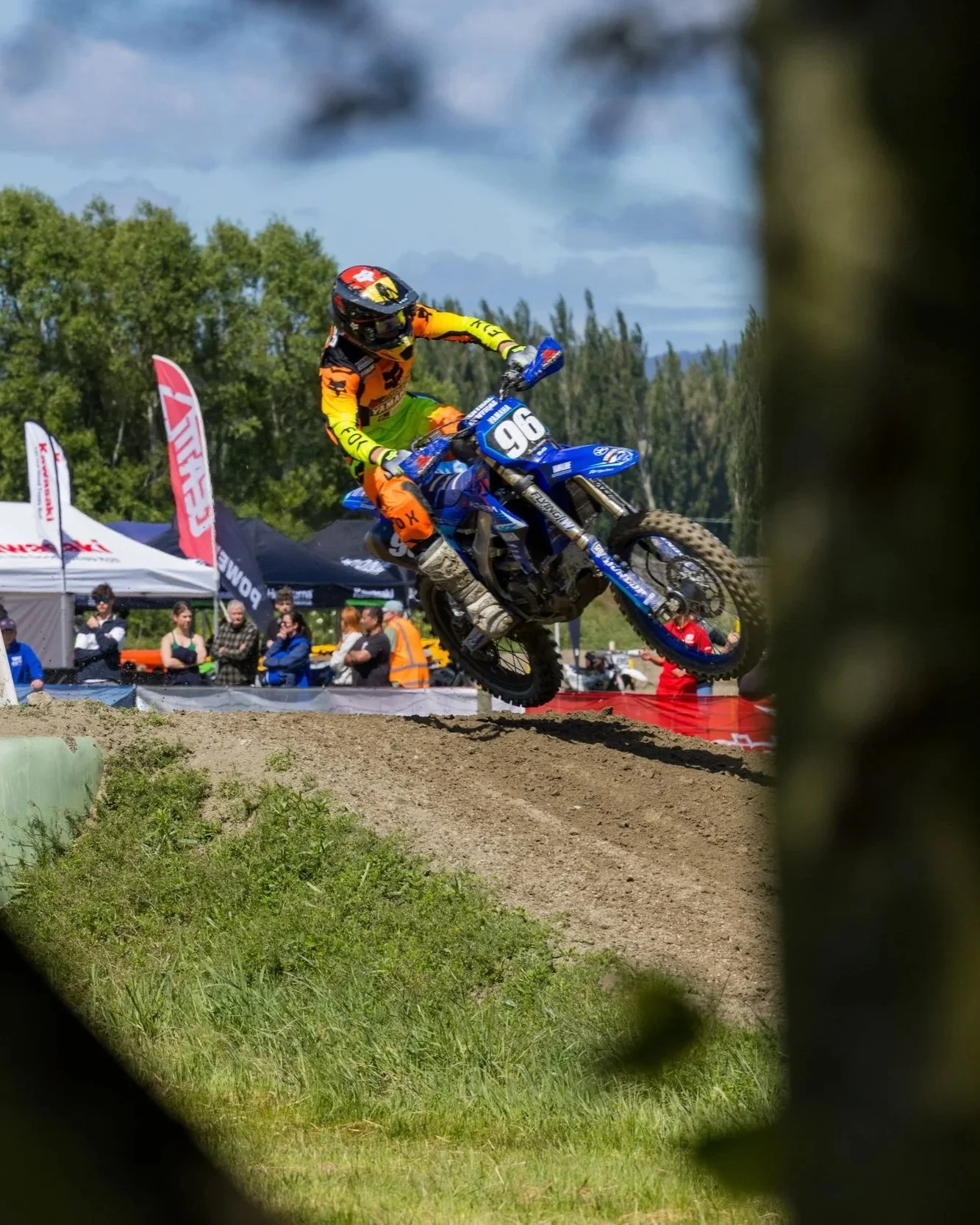 A motocross rider in orange and yellow gear riding a blue dirt bike, airborne over a dirt track, with spectators and tents in the background, viewed through a blurred frame.