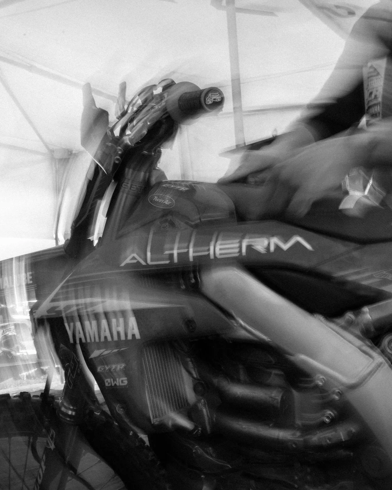 Black and white photo of a Yamaha motorcycle with 'ALCHEMY' written on the fuel tank, parked inside a room with a white ceiling and walls. A person is adjusting or working on the motorcycle, with their arm and hand visible.