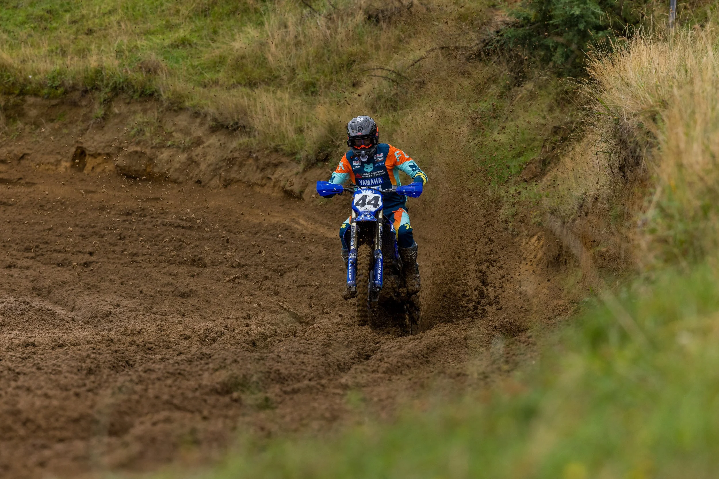 Motocross rider wearing a helmet and protective gear riding a dirt bike on a rough trail through grassy terrain.