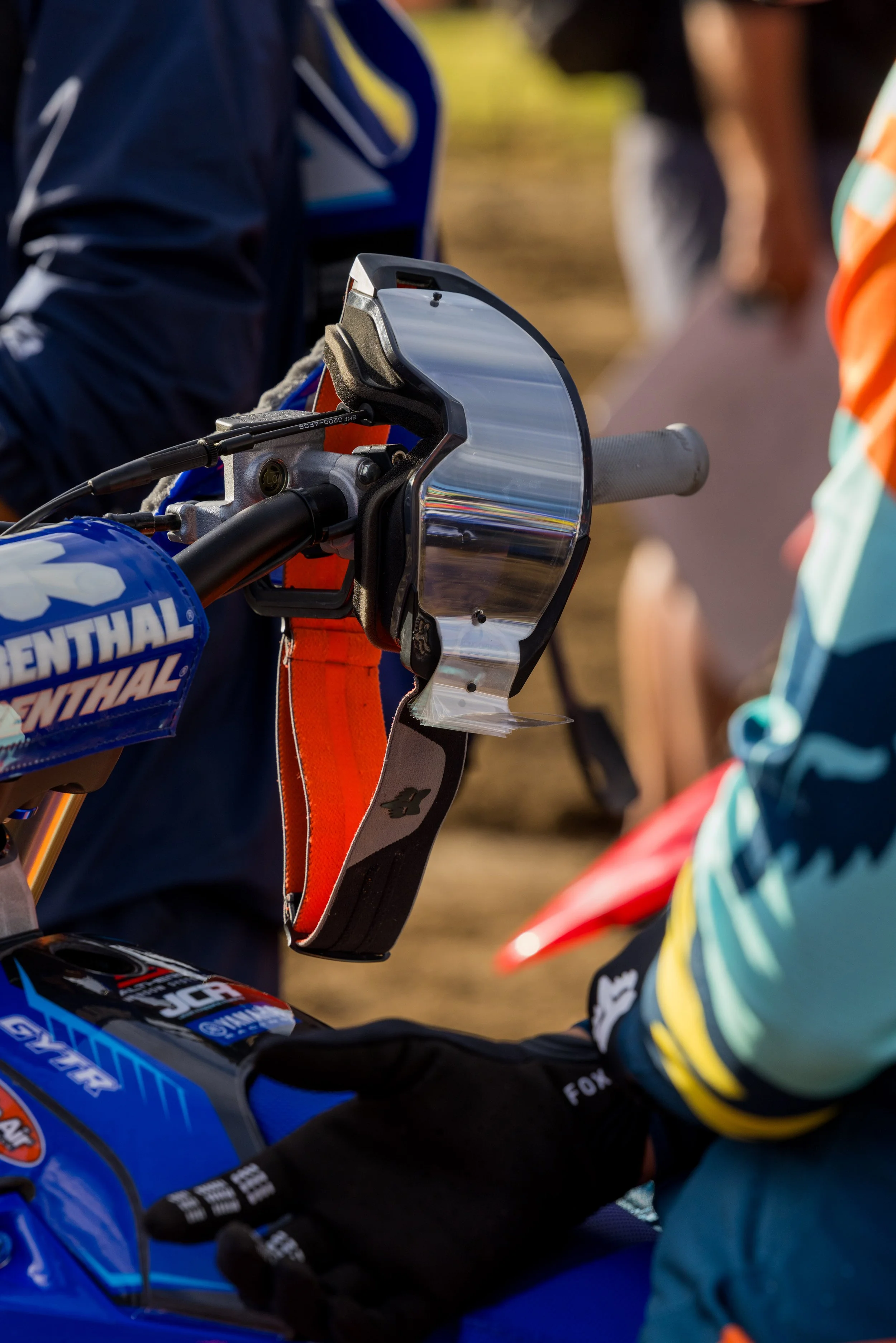 Close-up of a motorcycle handlebar with a tinted windshield, blue and orange gear, and a person's hand wearing a black glove, in a racing or off-road environment.