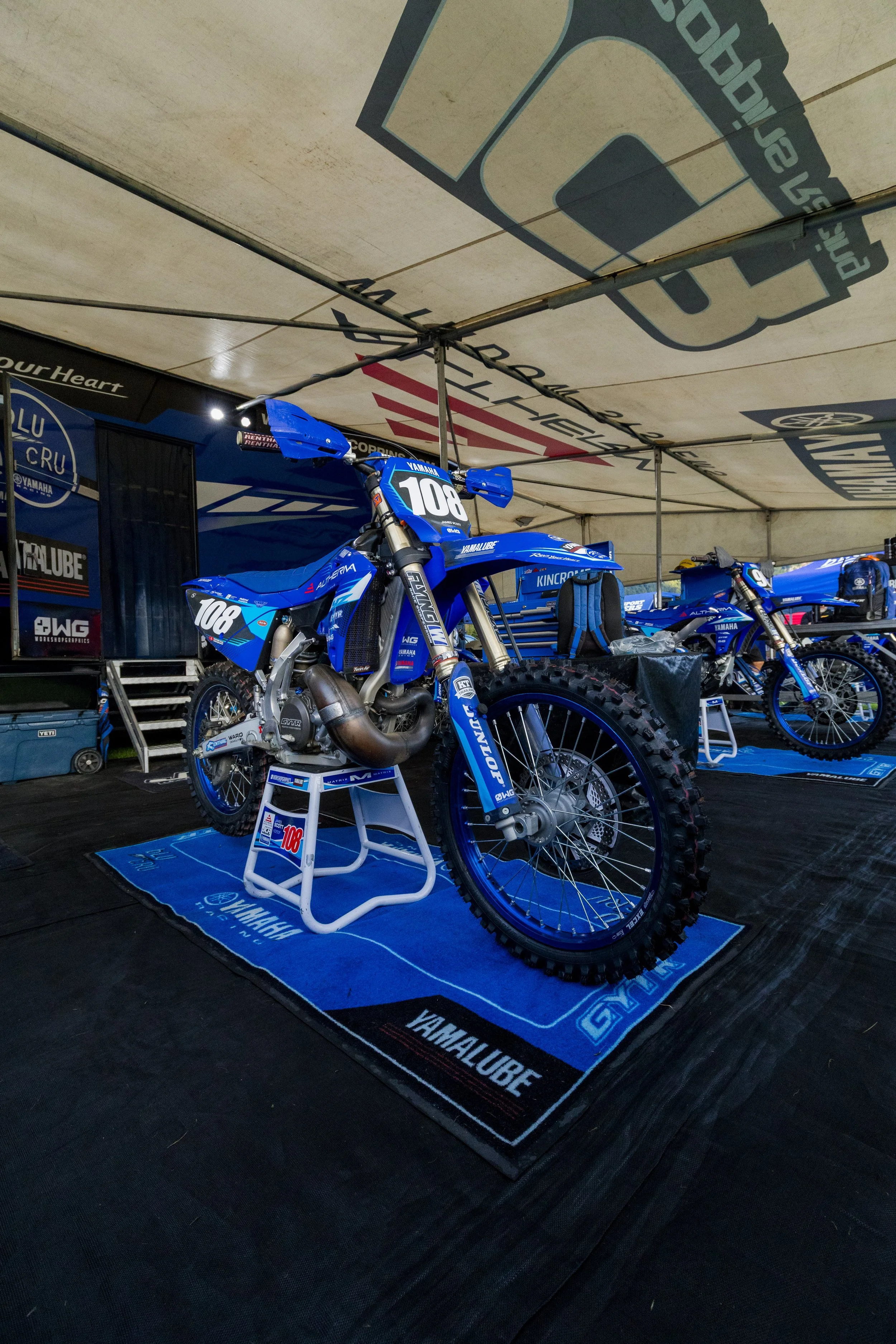 A blue Yamaha dirt bike with the number 108 on the front, displayed inside a fair tent on a Yamaha-branded mat, with storage containers and tools in the background.