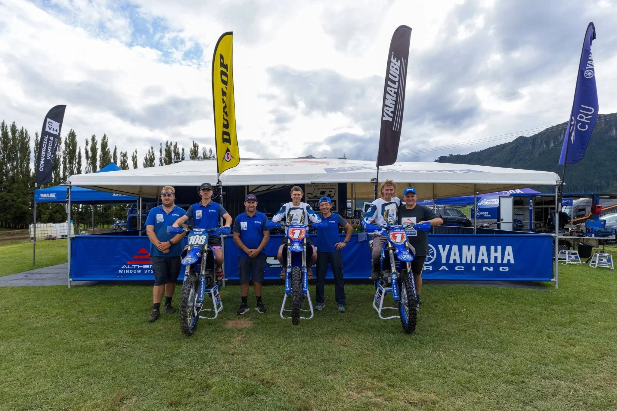 Yamaha motocross team posing with bikes and staff in front of a Yamaha racing tent outdoors, with flags and mountains in the background.