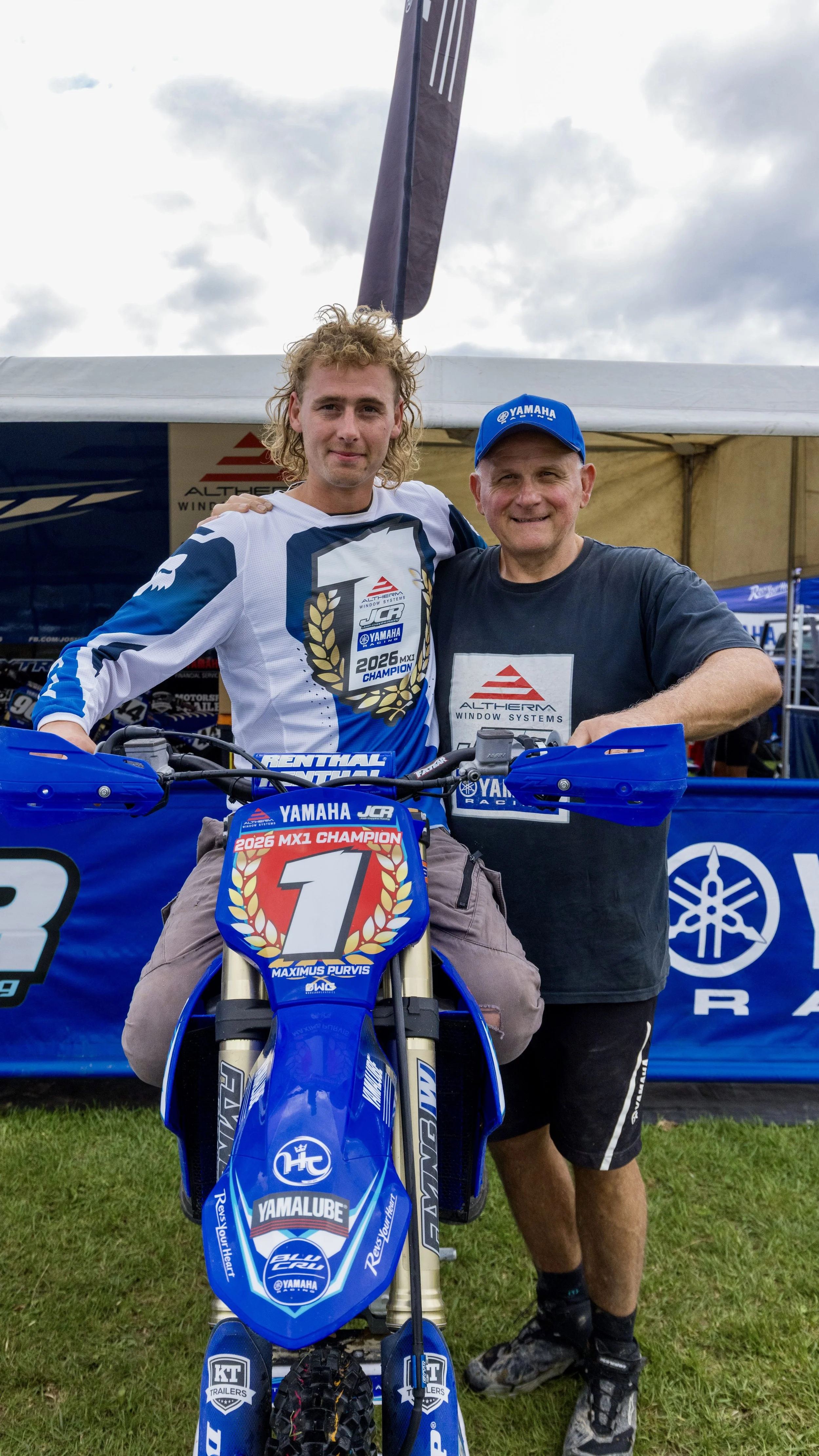 Two men at a motocross event, one sitting on a Yamaha dirt bike with a winner's plate, and the other standing beside him with his arm around his shoulder, at an outdoor event with tents and banners in the background.