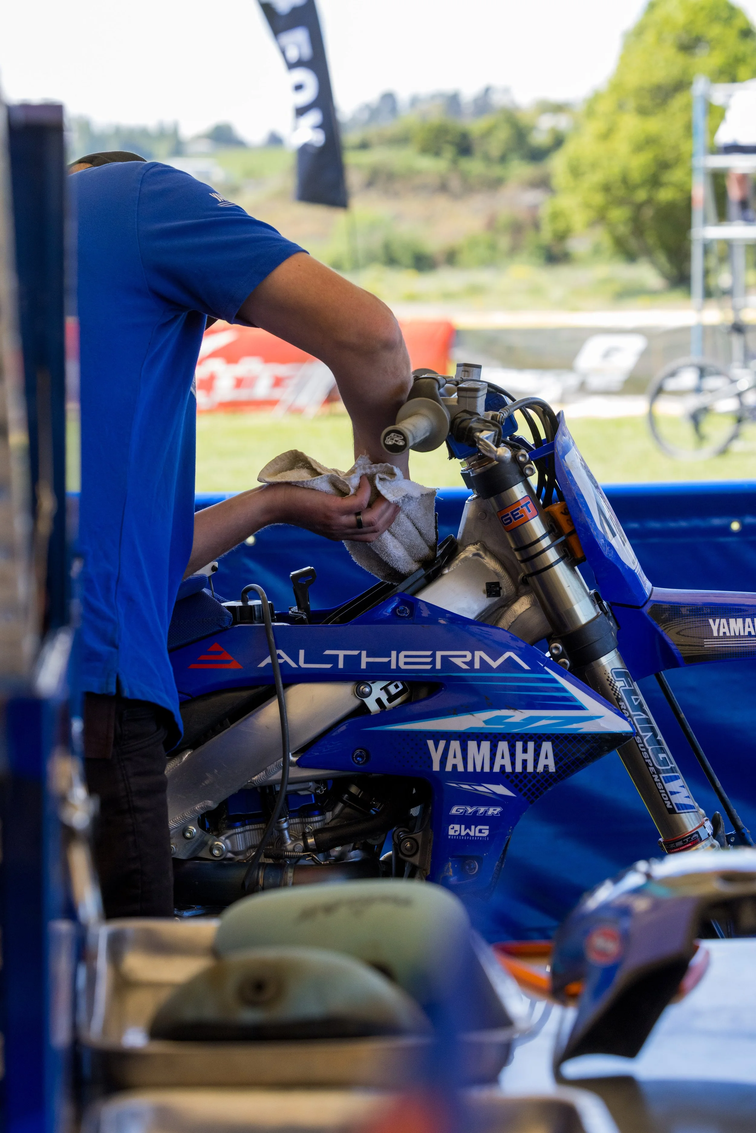 A motorcyclist working on a Yamaha motocross bike, wiping the handlebar area with a cloth, in an outdoor setting.