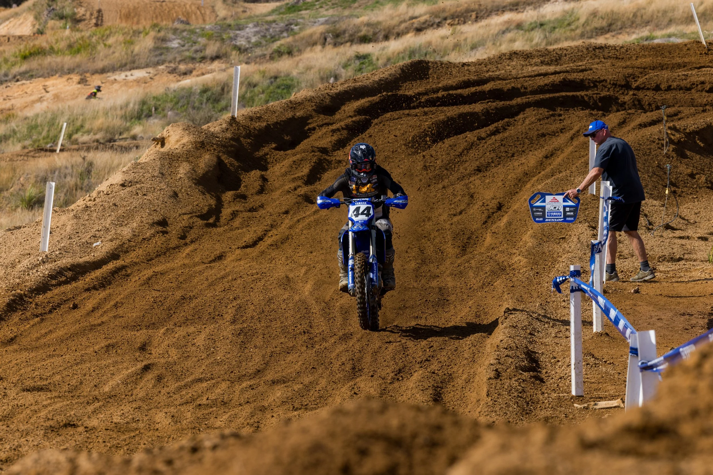 A motocross rider wearing a black helmet, black and blue gear, and riding a blue Yamaha motorcycle with the number 44 on it navigates a dirt track. A person in a black shirt and blue cap is managing the track, while another rider in the distance is o