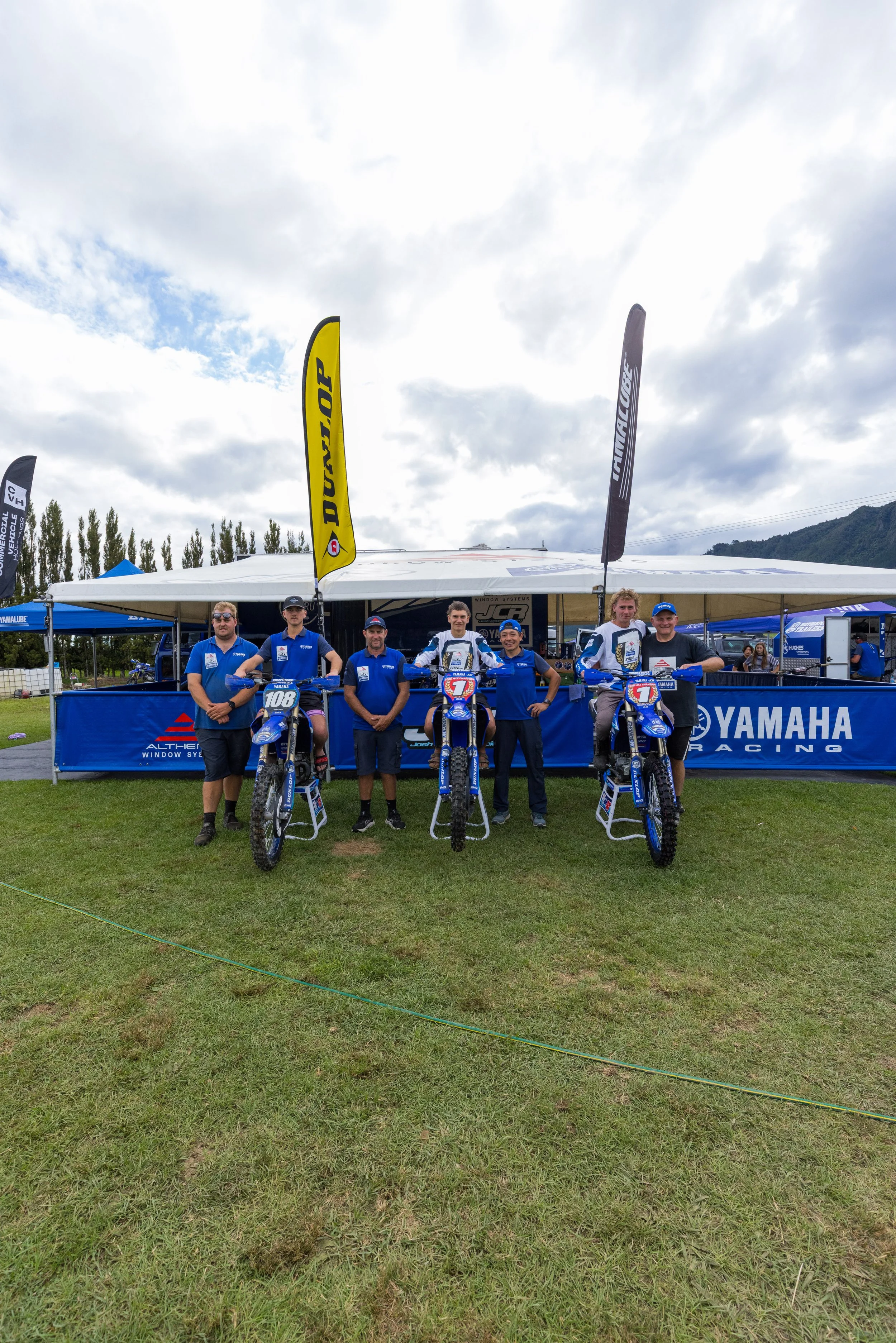 A group of six people standing in front of three Yamaha racing dirt bikes at an outdoor racing event, with a large Yamaha banner behind them and paper flags with sponsor logos on the poles. The sky is cloudy with some blue sky peeking through, and there are tents and trees in the background.