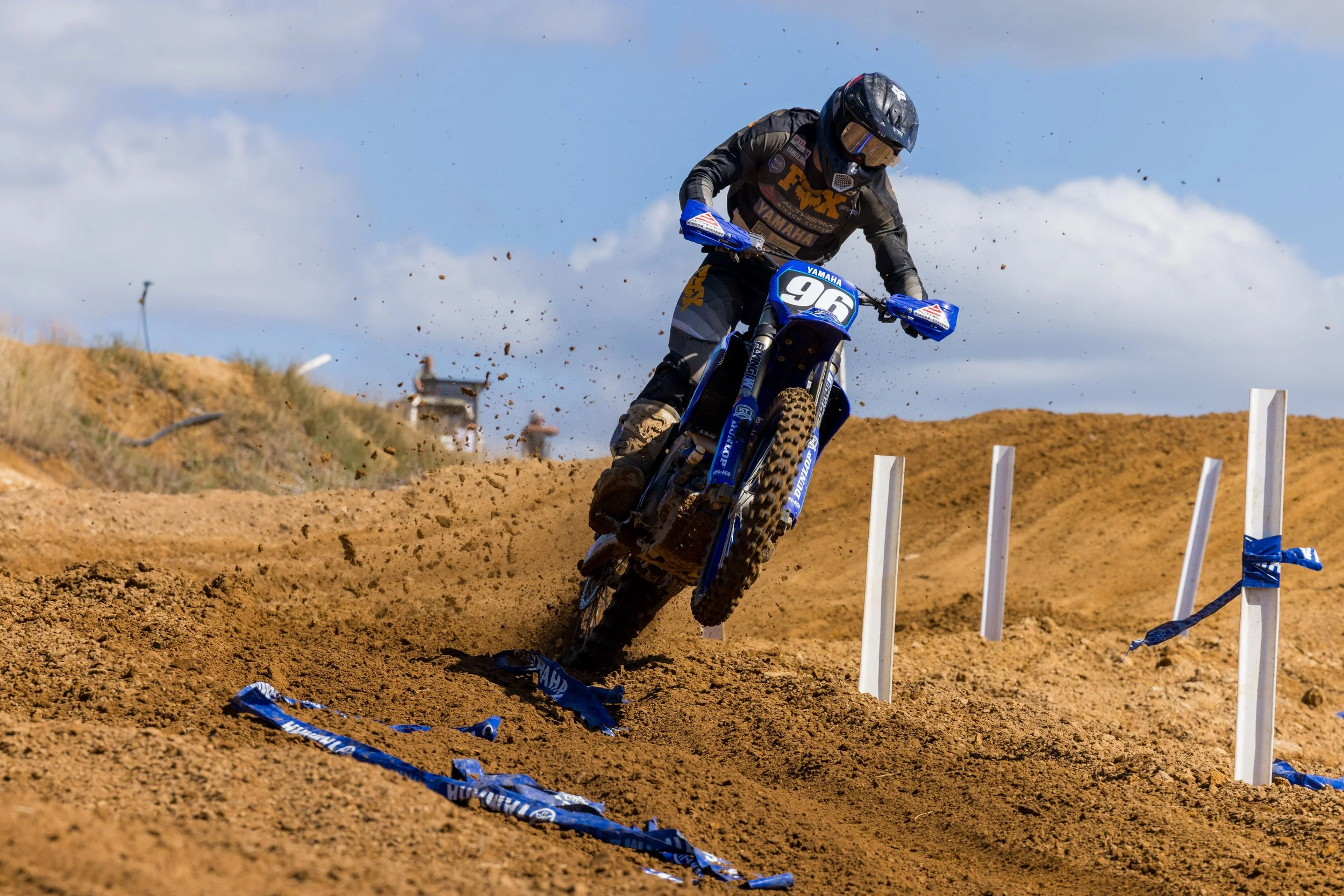 A motocross rider in gear riding a blue Yamaha dirt bike on a dirt track with dirt flying and blue sky with clouds in the background.