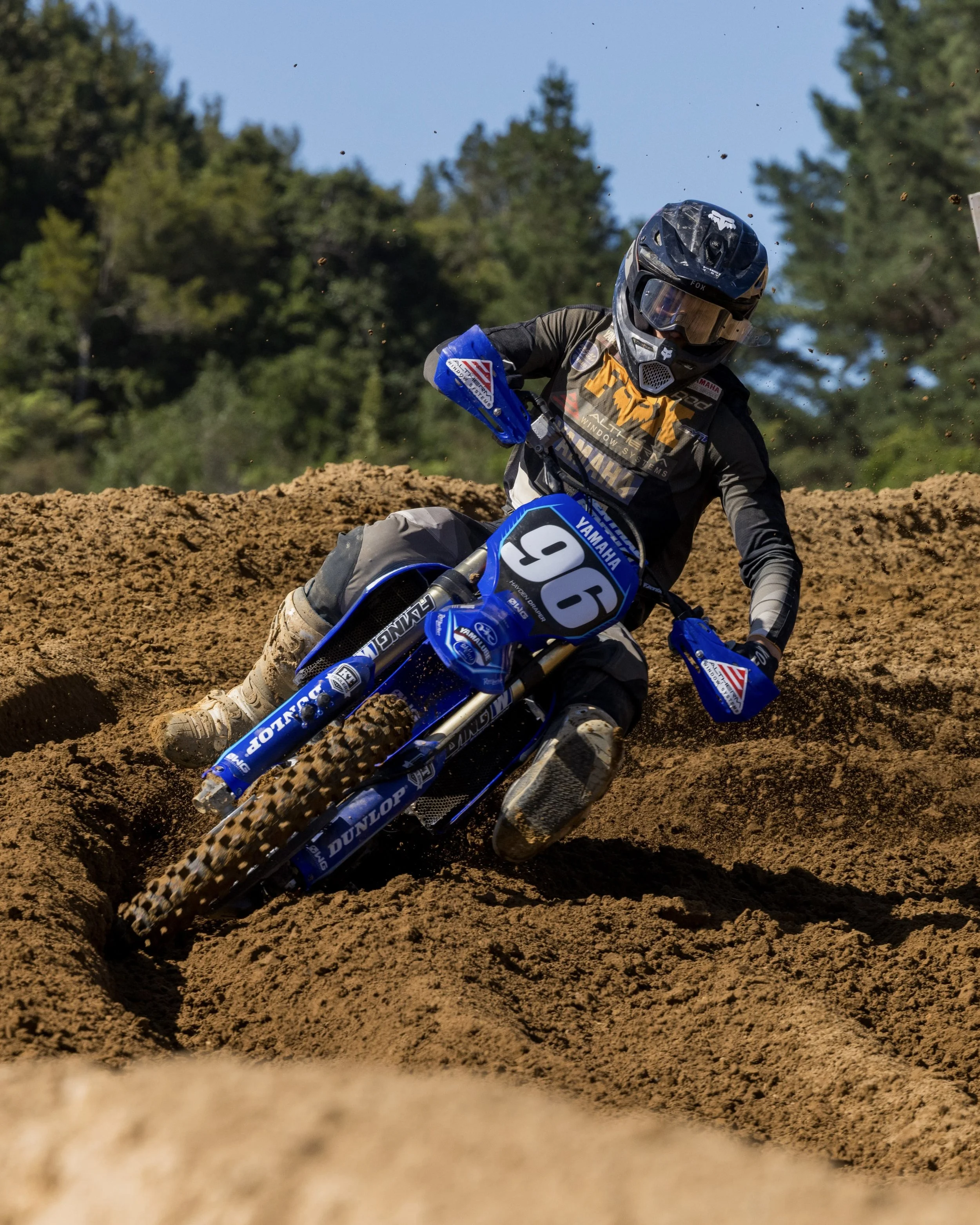 Motocross rider on a dirt track during daytime, wearing black helmet and gear, riding a blue Yamaha motorcycle, kicking up dirt with trees in the background.