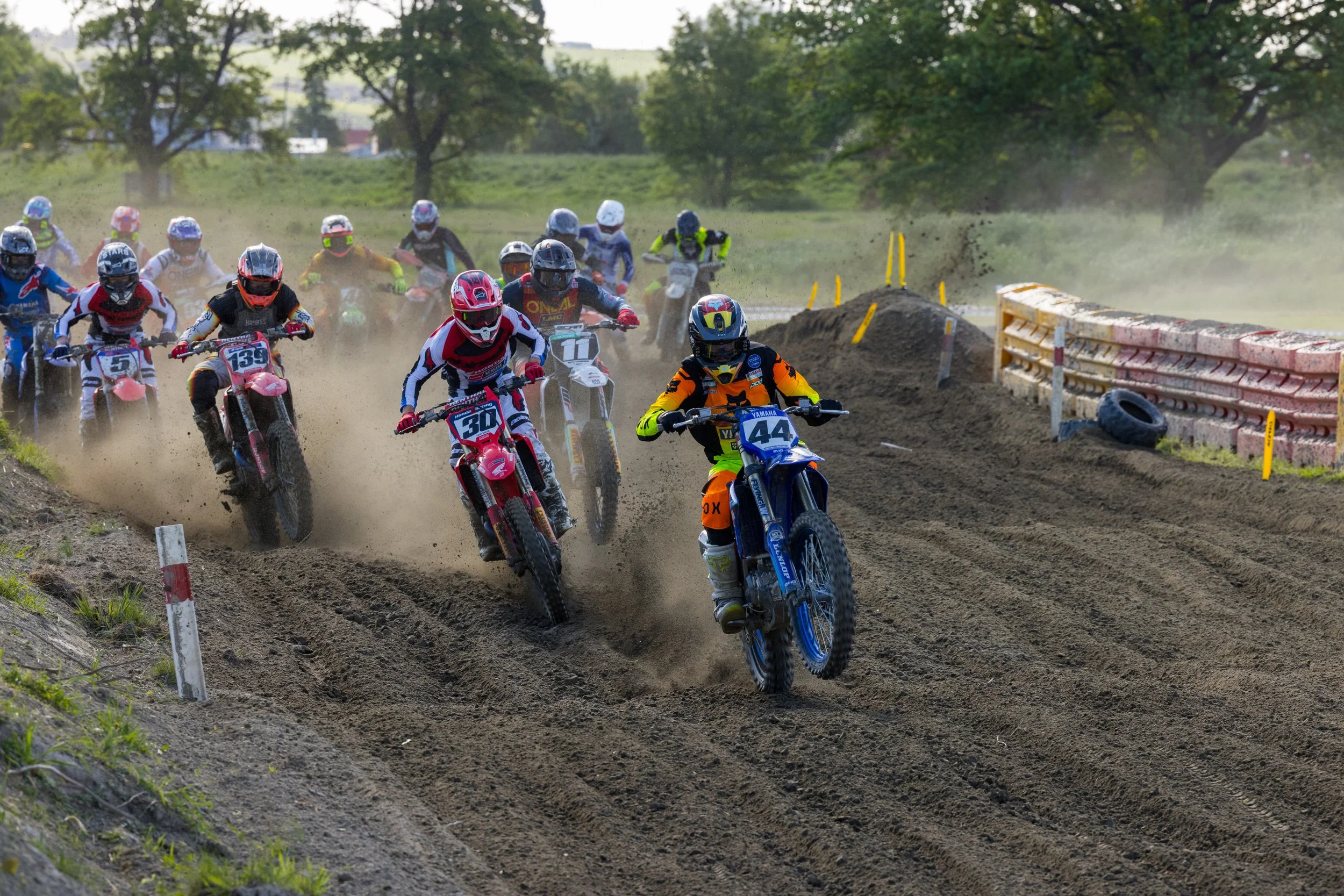 A group of motocross riders racing on a dirt track, kicking up dust, with trees and a fence in the background.