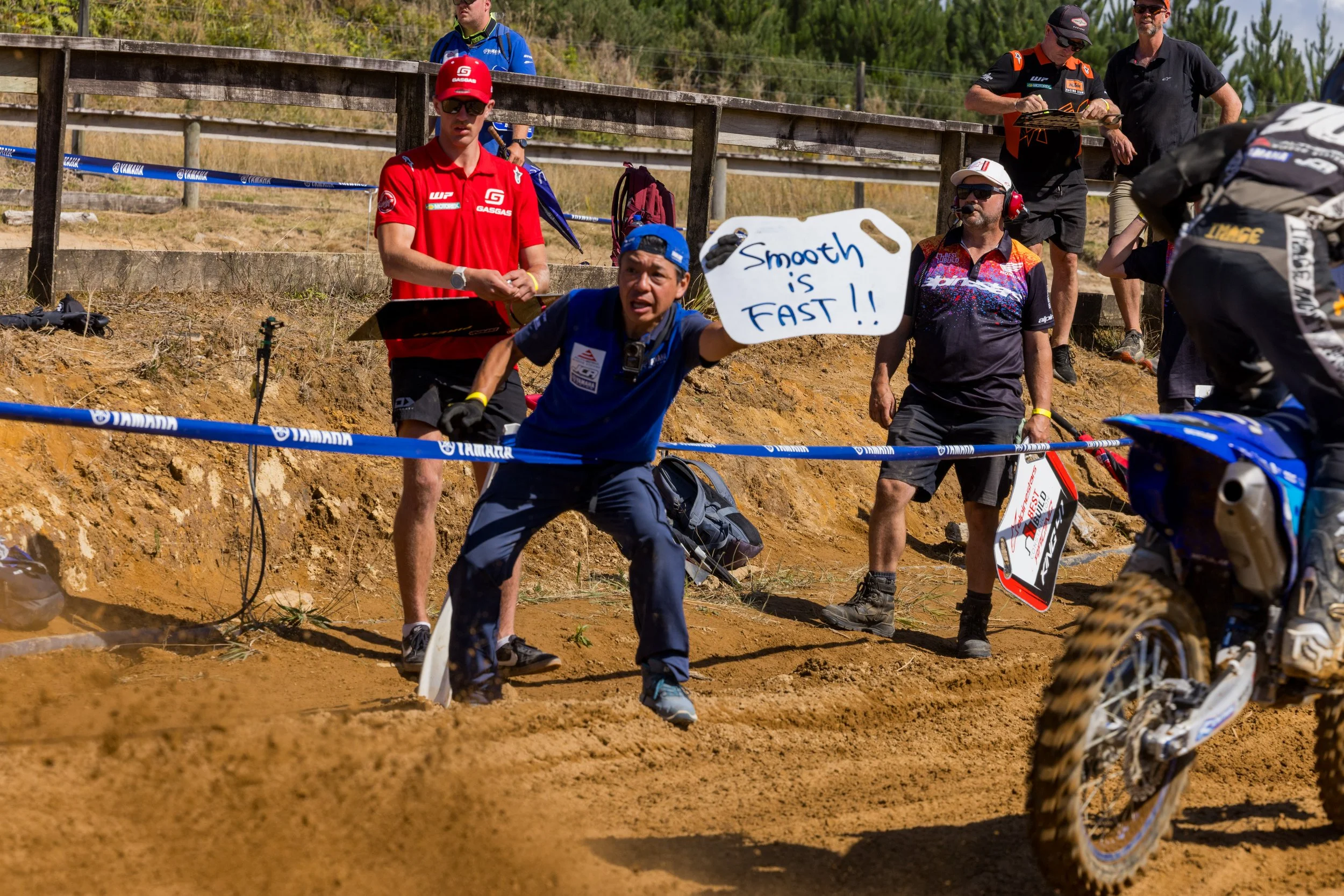 A group of people at a dirt bike race, with some seated and others standing. One person is holding a sign that says 'Smooth is FAST!!', and a rider on a dirt bike is partially visible in the foreground.