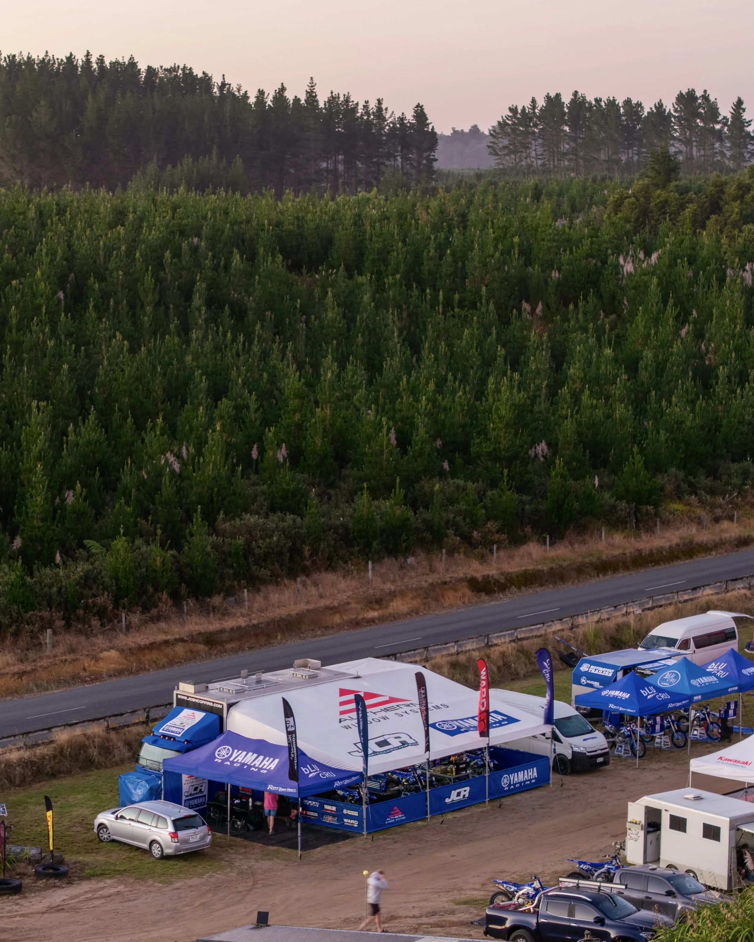 A racing event setup with tents and vehicles near a forested area, with scooter motorcycles parked nearby and people walking around.