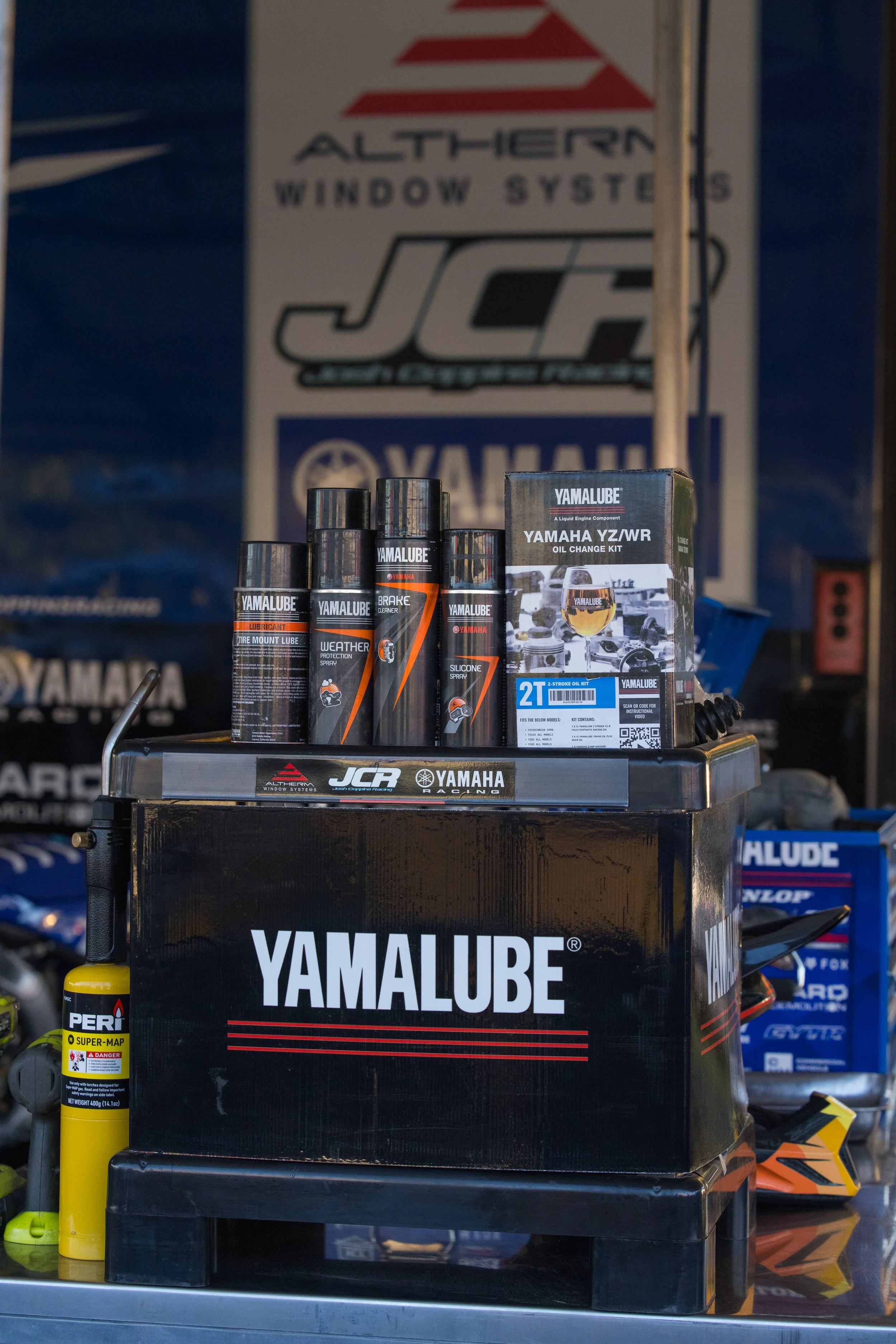 Yamalube motor oil products including oil change kit, brake cleaner spray, weather protection spray, and silicone spray displayed on a black stand with Yamaha and JCR logos. In the background, there is a sign with the Altherm Window Systems logo and 